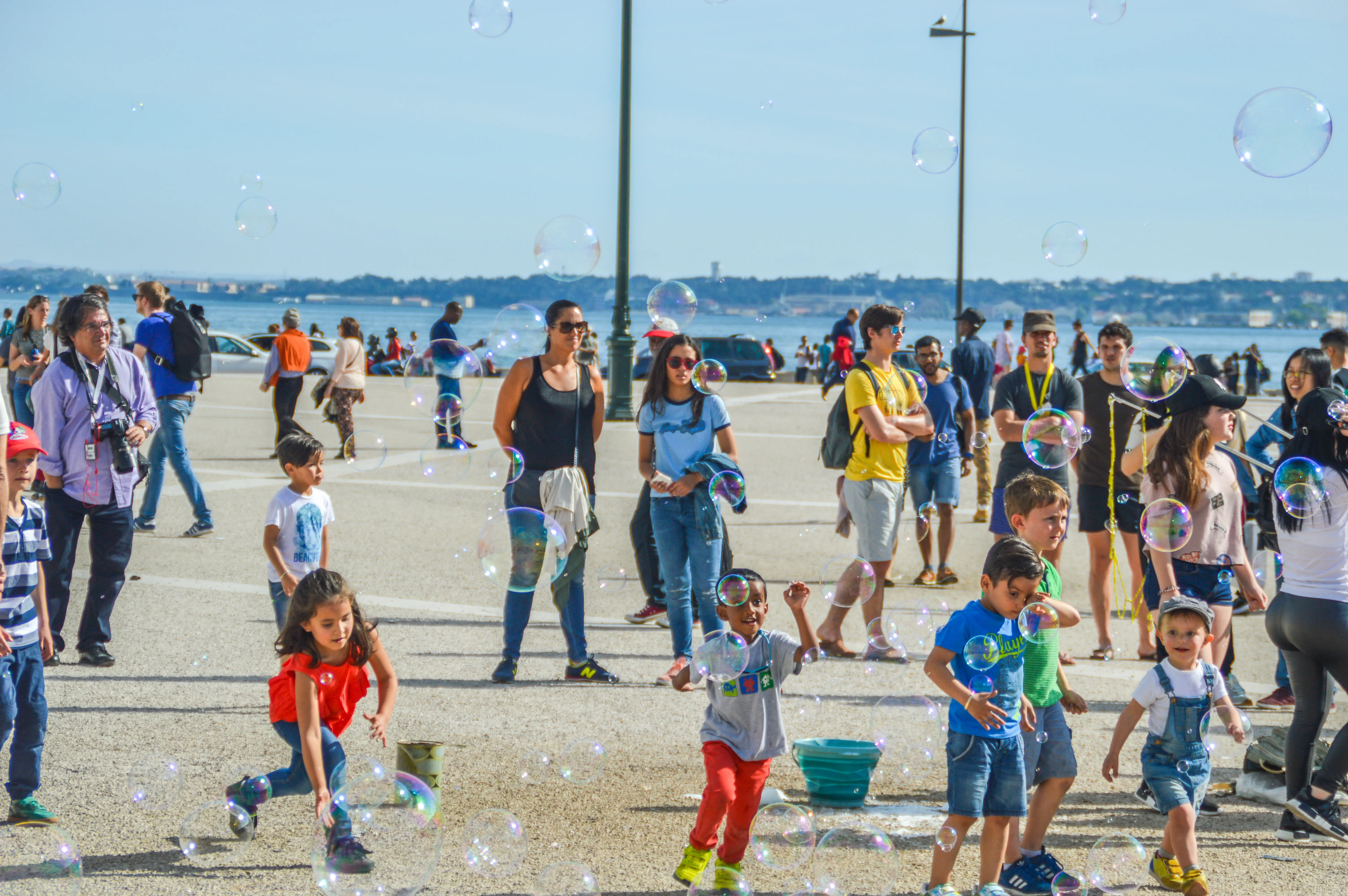 A diverse group of people, including children and adults, are playing with large, iridescent soap bubbles in an outdoor area near a body of water. The sky is blue and clear, and the sun is shining.