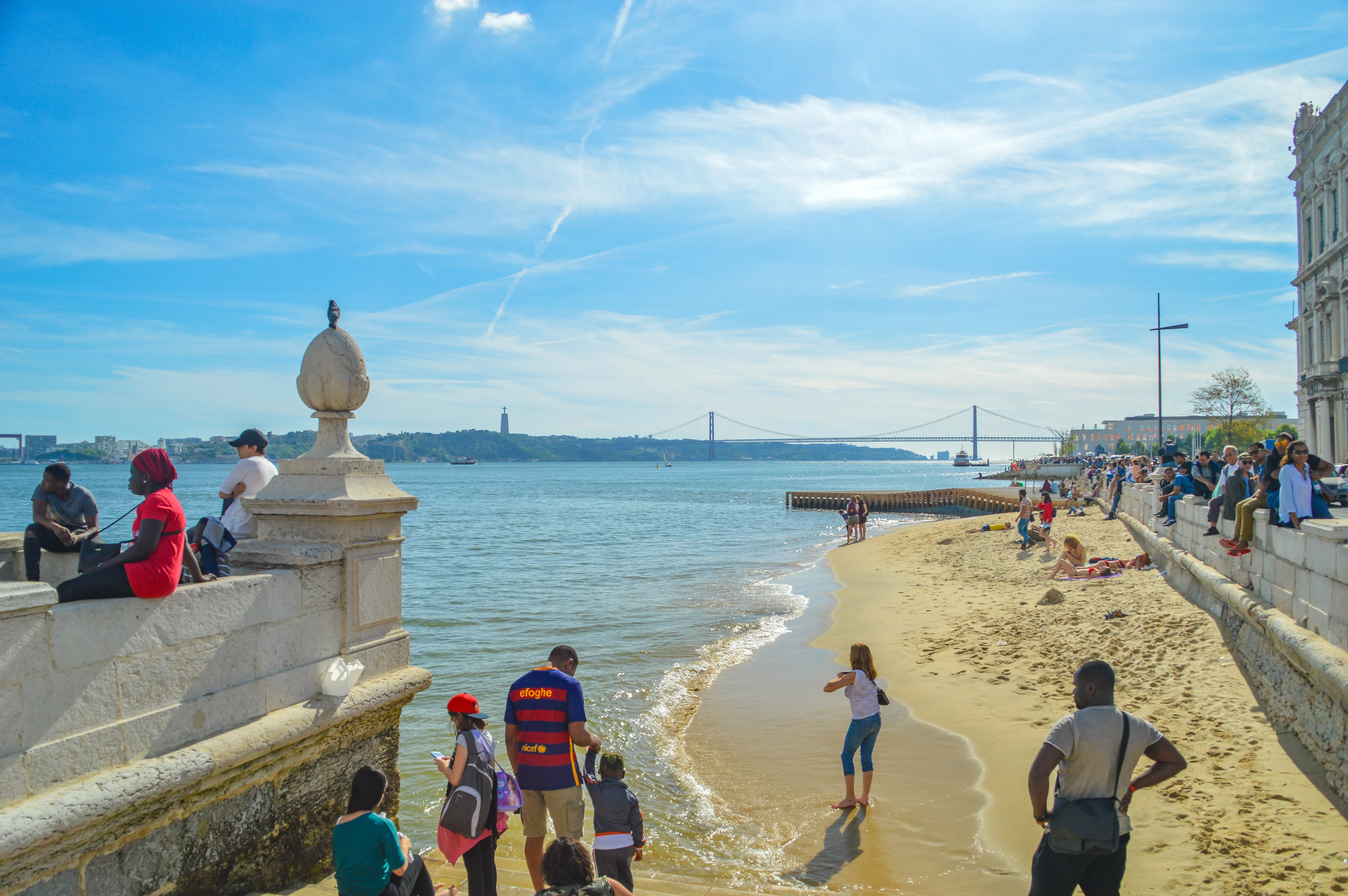 A sunny day at the Lisbon waterfront, with the 25 de Abril Bridge spanning the Tagus River and people enjoying the beach and promenade.