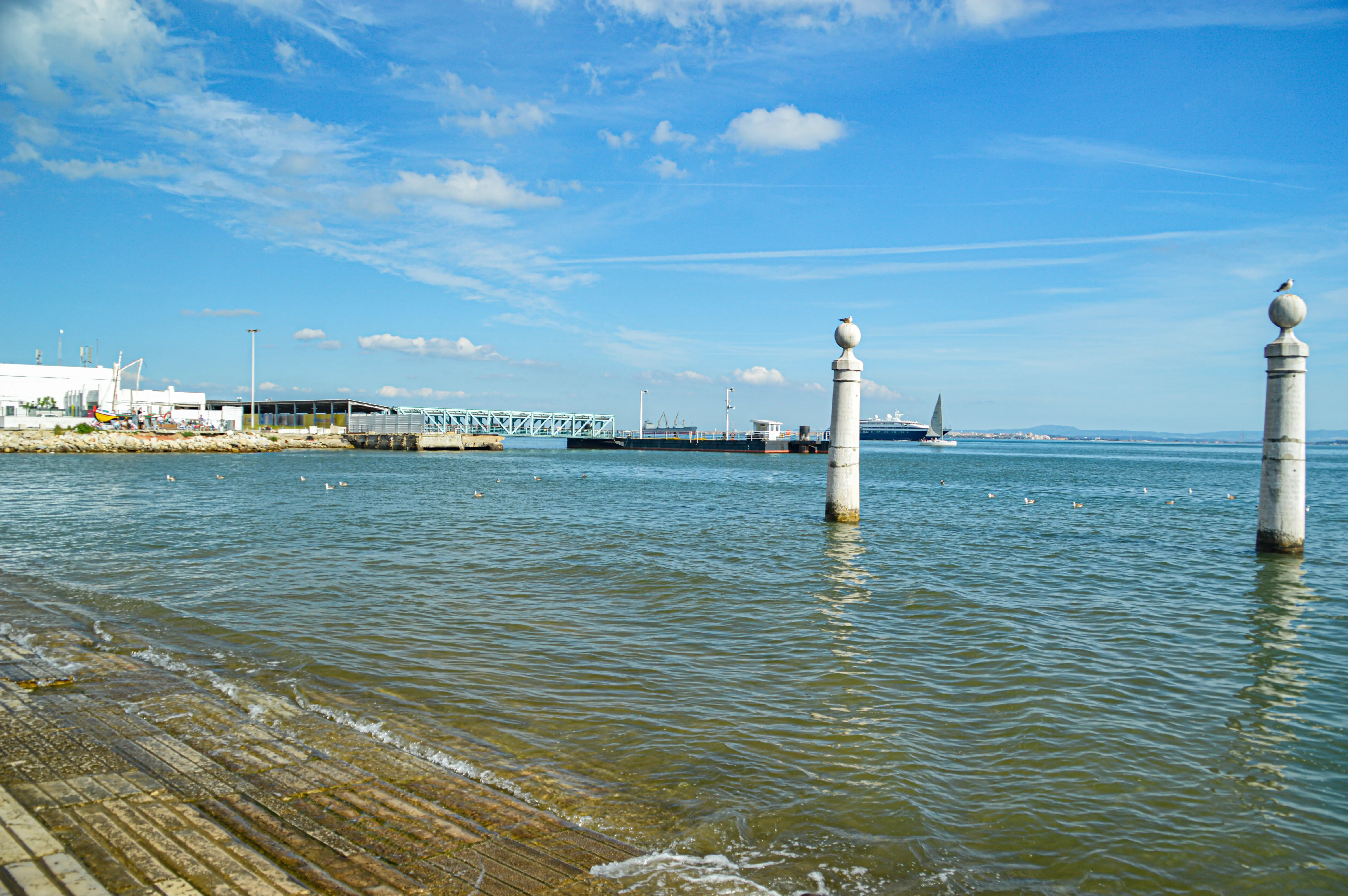 A wide shot of a waterfront scene showing two white stone pillars in the foreground. In the middle ground, a large cruise ship sails on the water, accompanied by a sailboat. A modern bridge and industrial buildings line the distant shore. Seagulls are visible on the water.