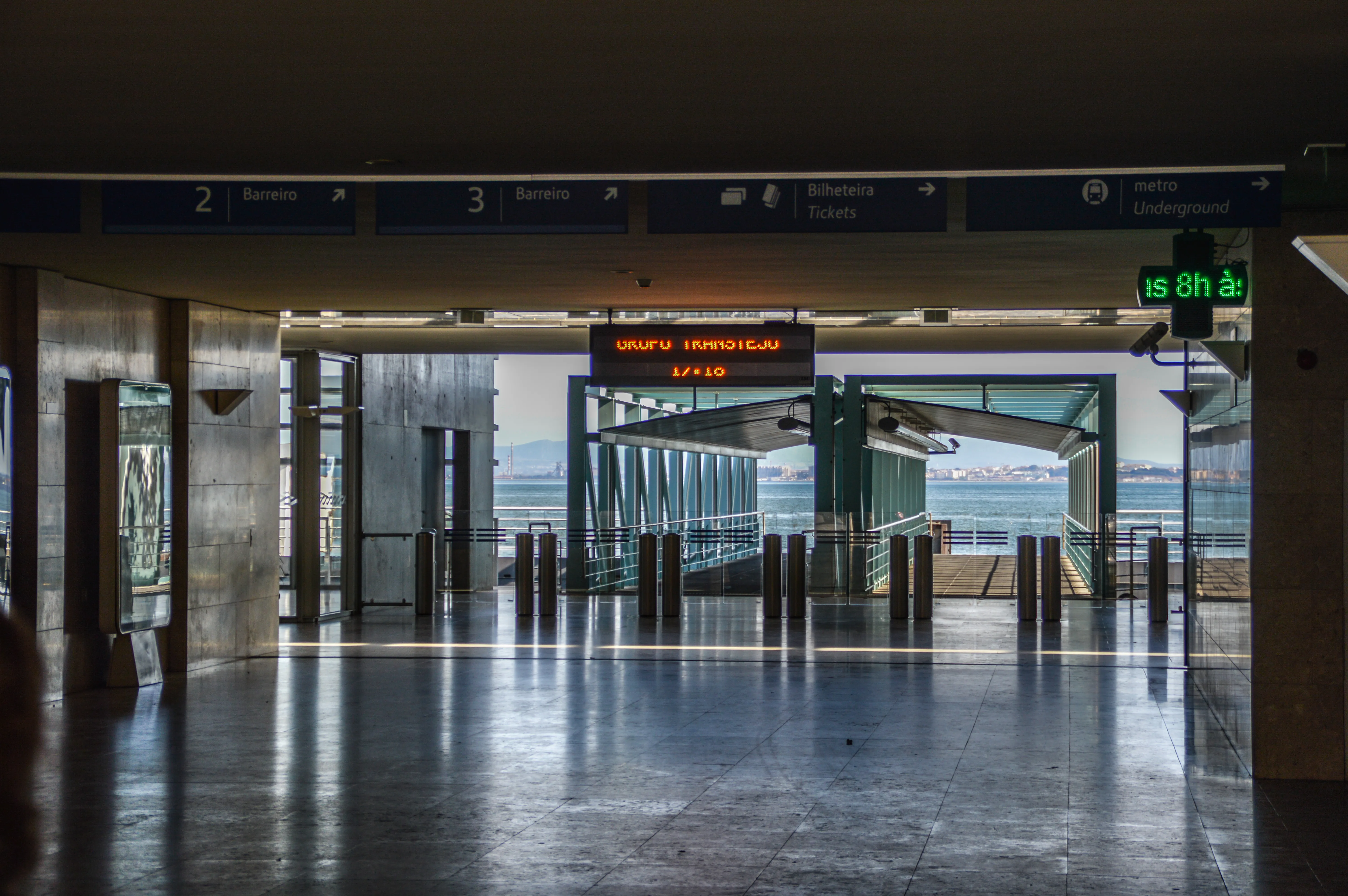 A woman stands near ticket barriers in a well-lit transport station with directional signs indicating platforms, tickets, and the metro.