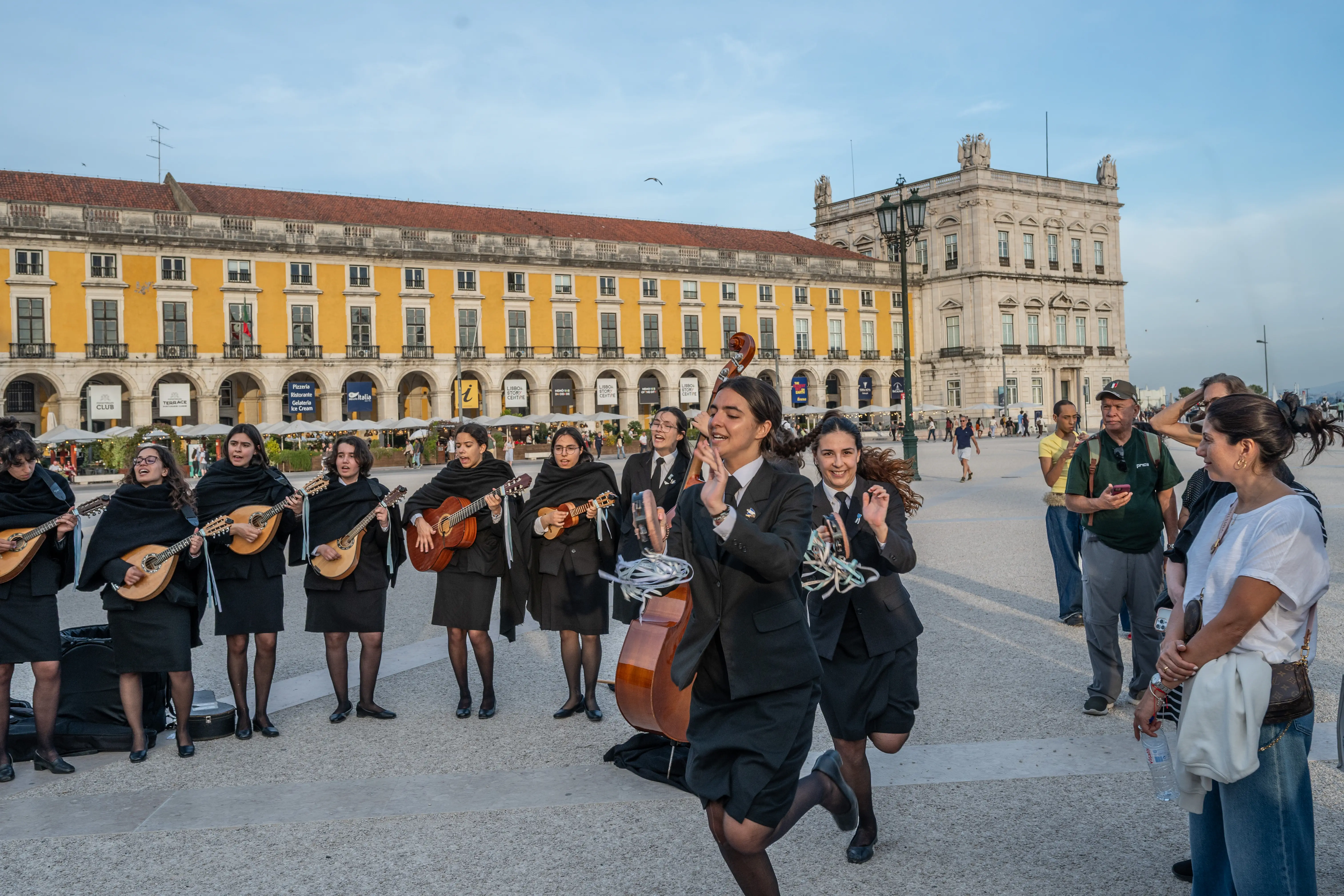 A group of young women in black uniforms and capes play stringed instruments and sing in Lisbon's Praça do Comércio, with yellow buildings and a stone palace in the background.