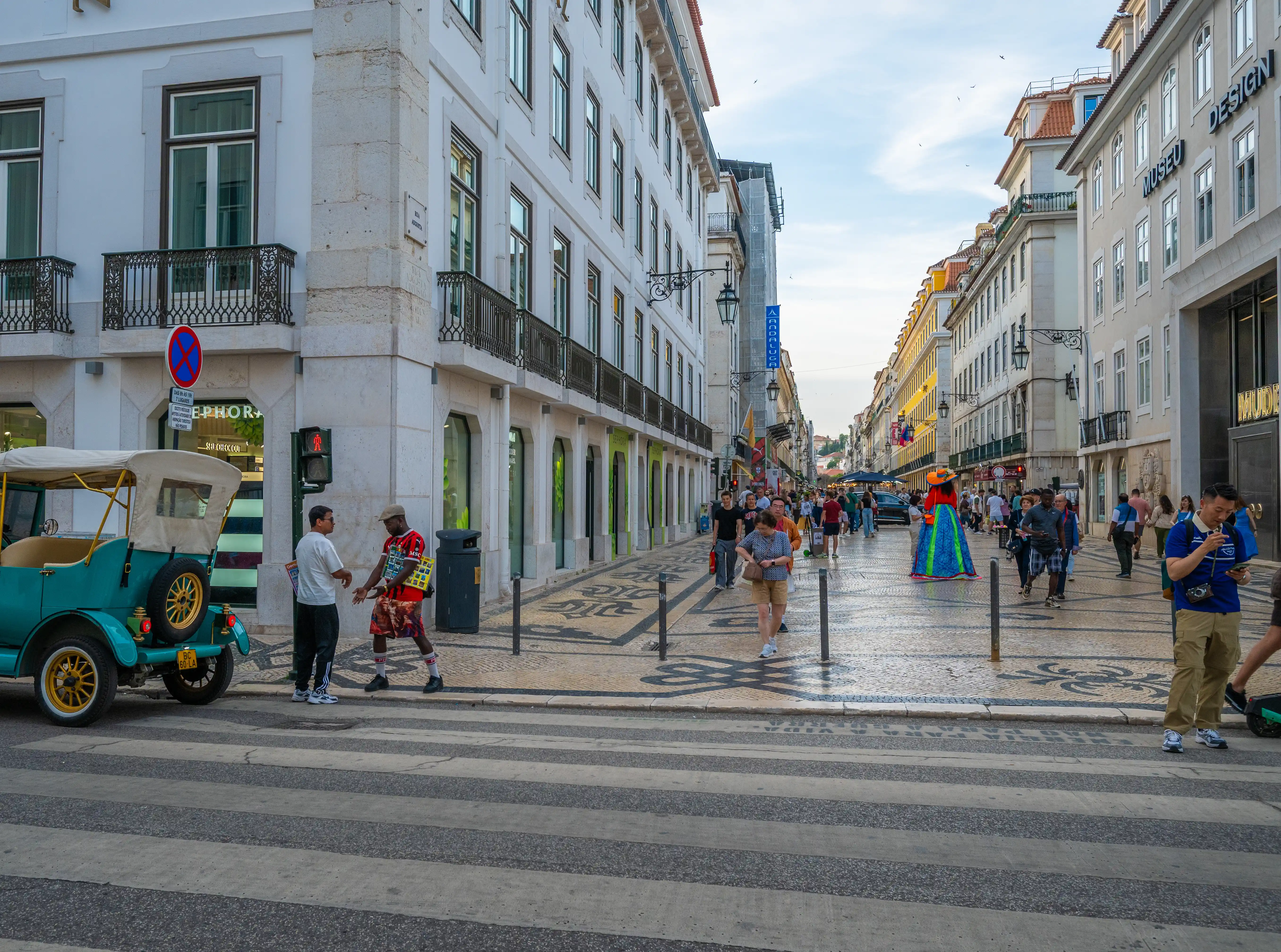 A turquoise vintage car at a pedestrian crossing on a busy Lisbon street, with people walking on the pavement and buildings on either side.