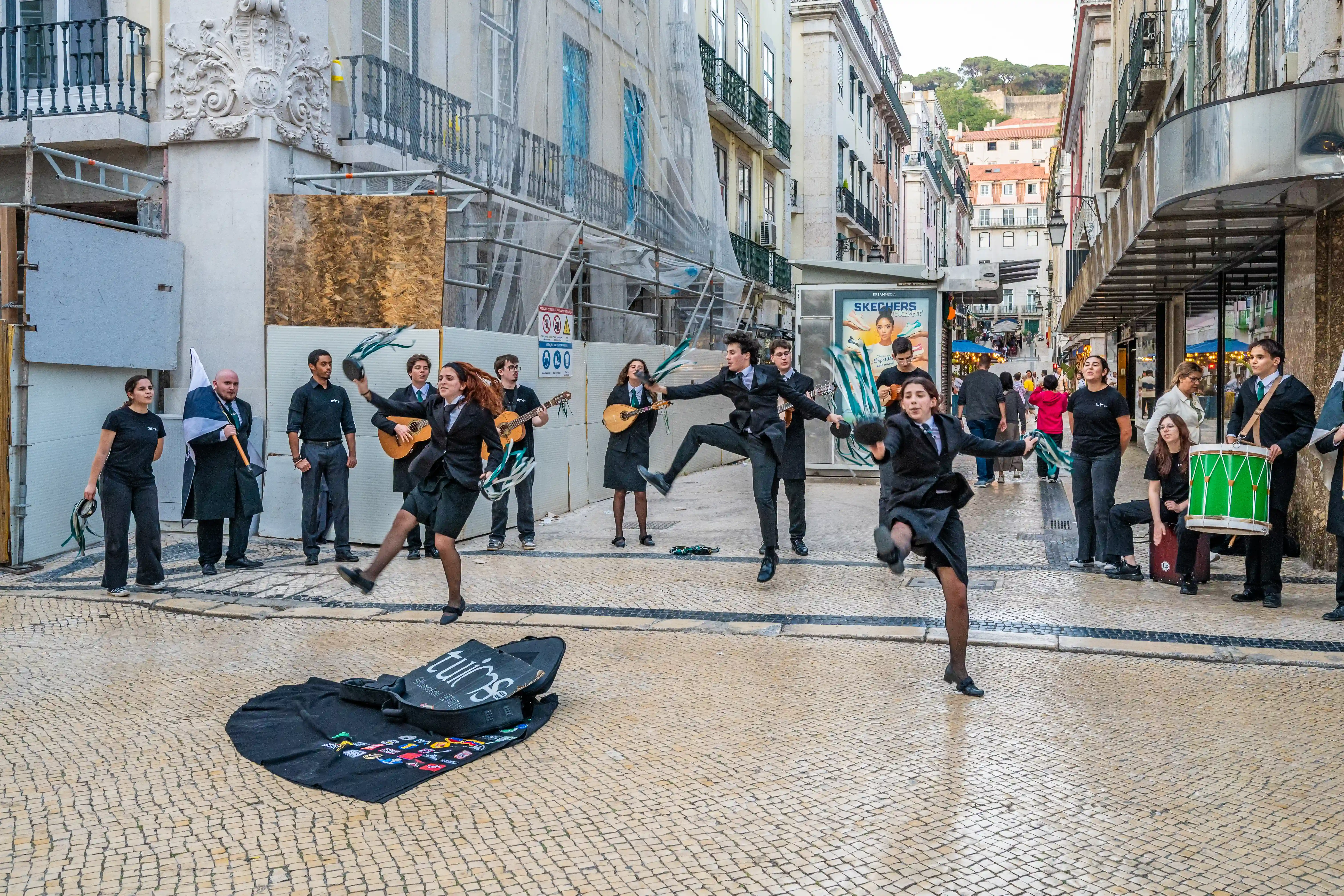 A street performance group in Lisbon, Portugal, with two female dancers in black uniforms performing in front of guitarists and a drummer.