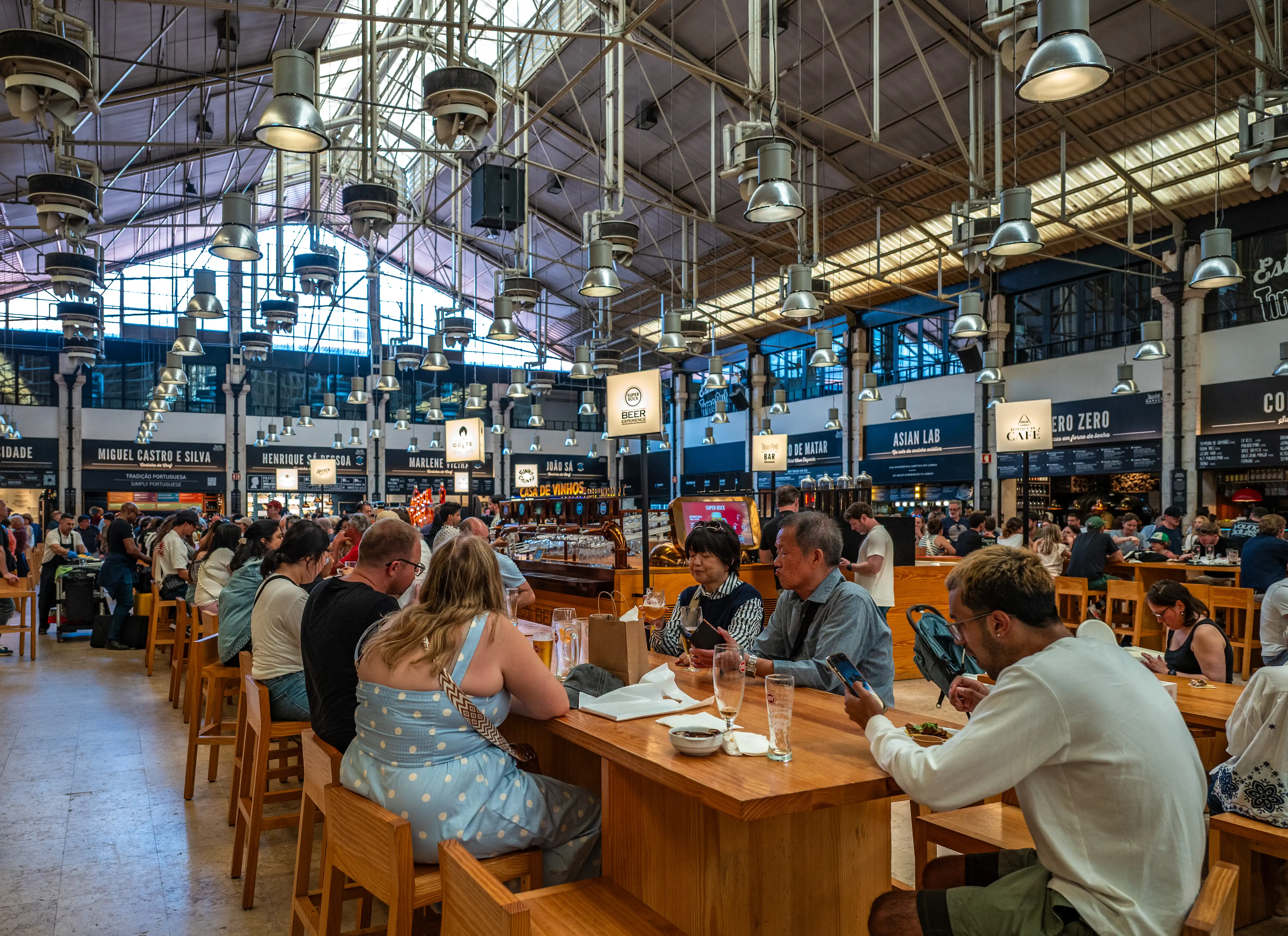 A wide shot of the interior of Time Out Market Lisboa, showing numerous people dining at wooden tables and benches, with a large Time Out Market Lisboa sign hanging from the ceiling. Various food stalls with signage are visible in the background.