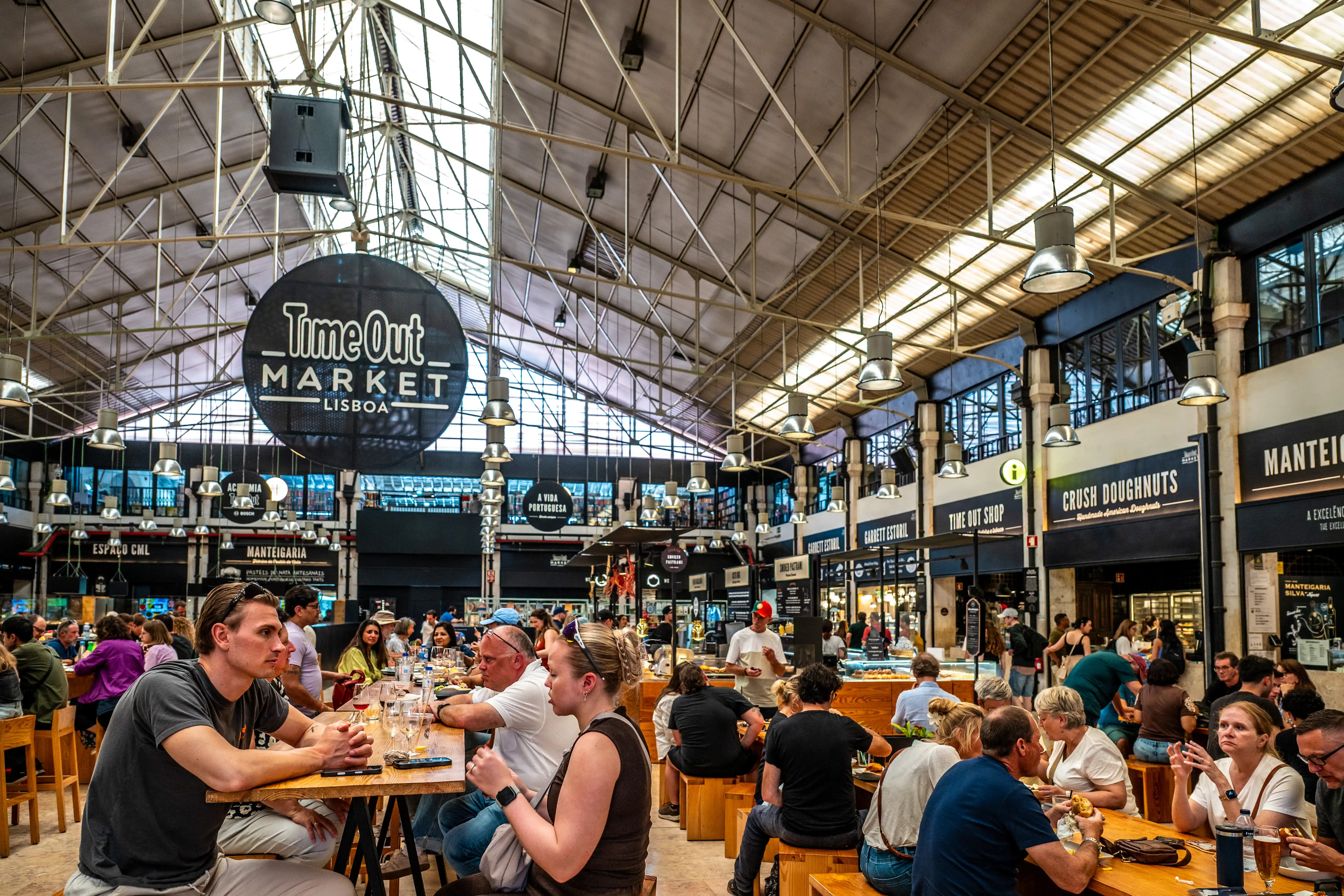 A busy interior view of Time Out Market Lisbon, showing diners at wooden tables in front of various food stalls under a high, industrial-style ceiling with hanging lights.