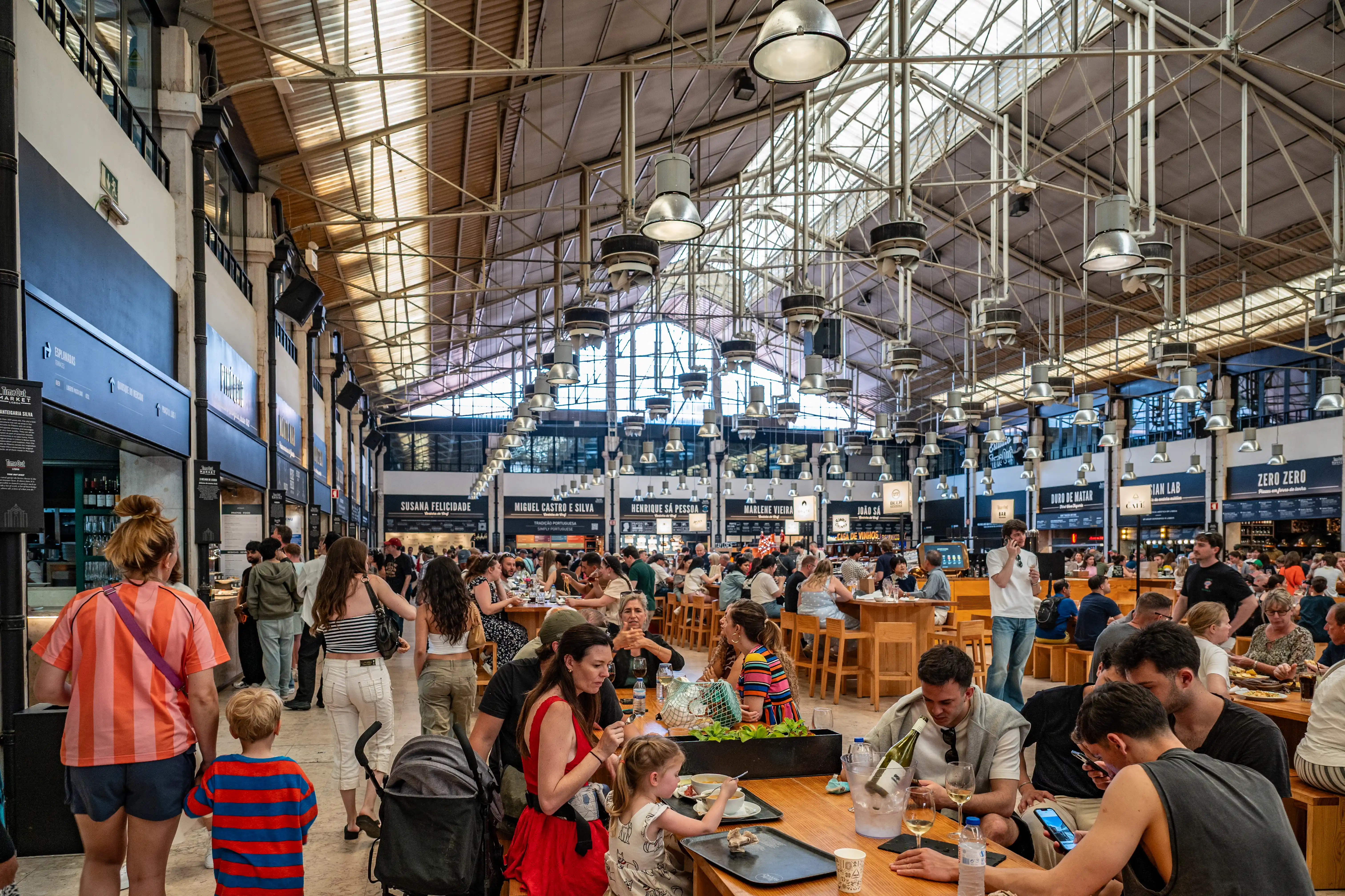 Interior view of a busy food market with people eating at tables and numerous food stalls.