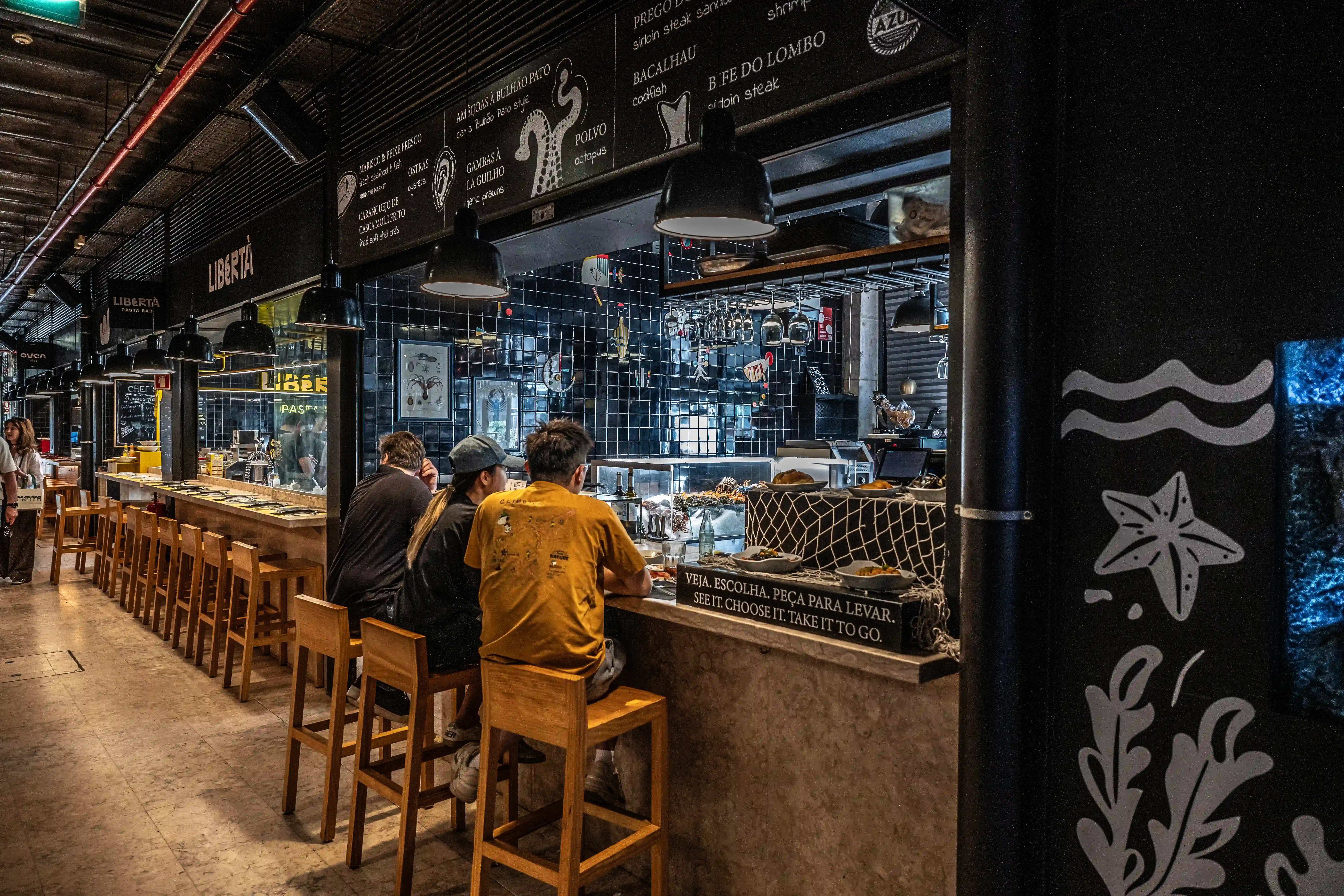 Three people sit at a counter in a food market. Behind them, a tiled wall displays artwork and menus. Wooden stools line the counter, and overhead lights illuminate the space.