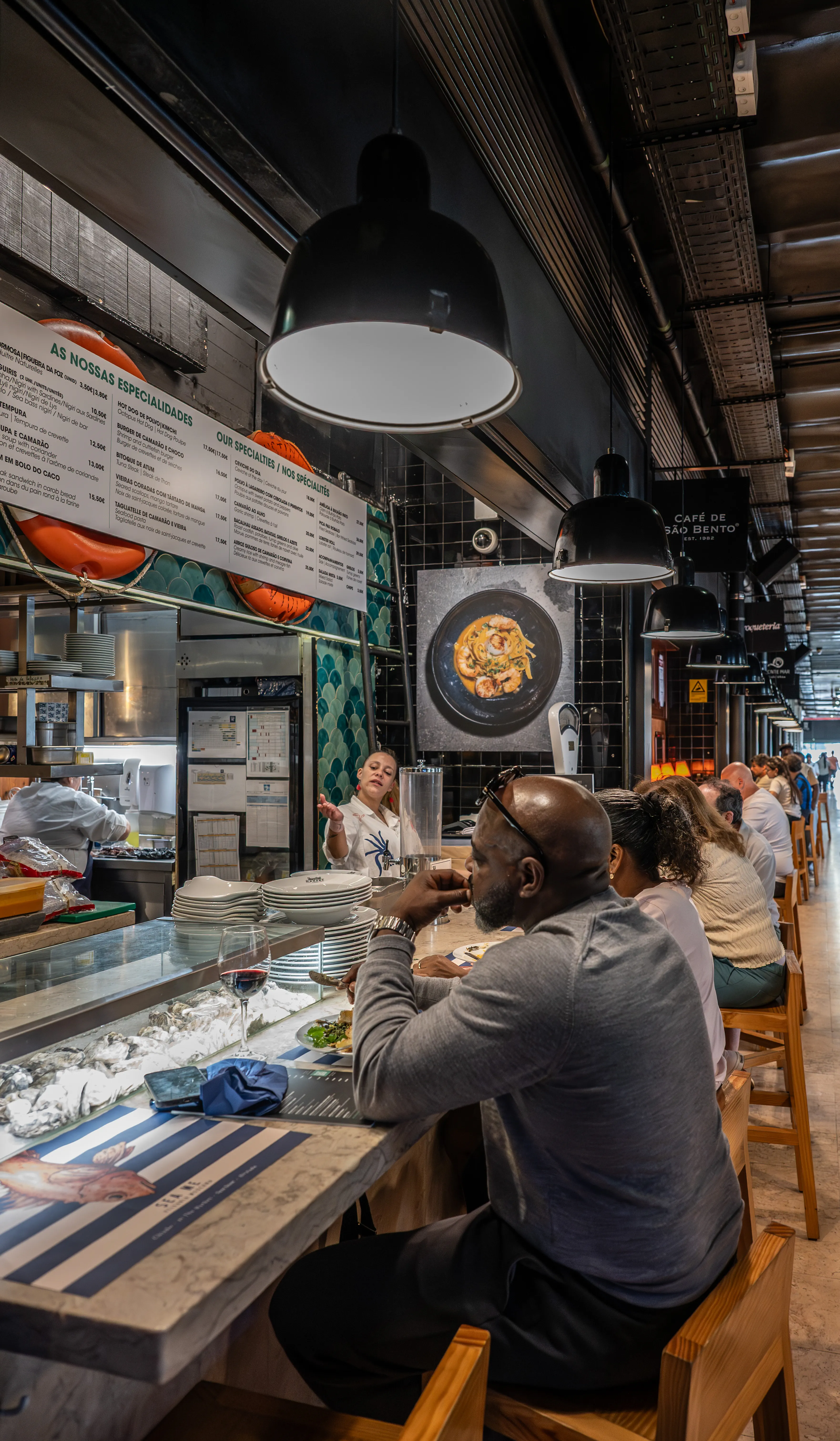 A diverse group of people are seated at a marble-topped counter in a busy indoor food market. A server in a white shirt is talking to customers, and there are menus and plates of food visible.