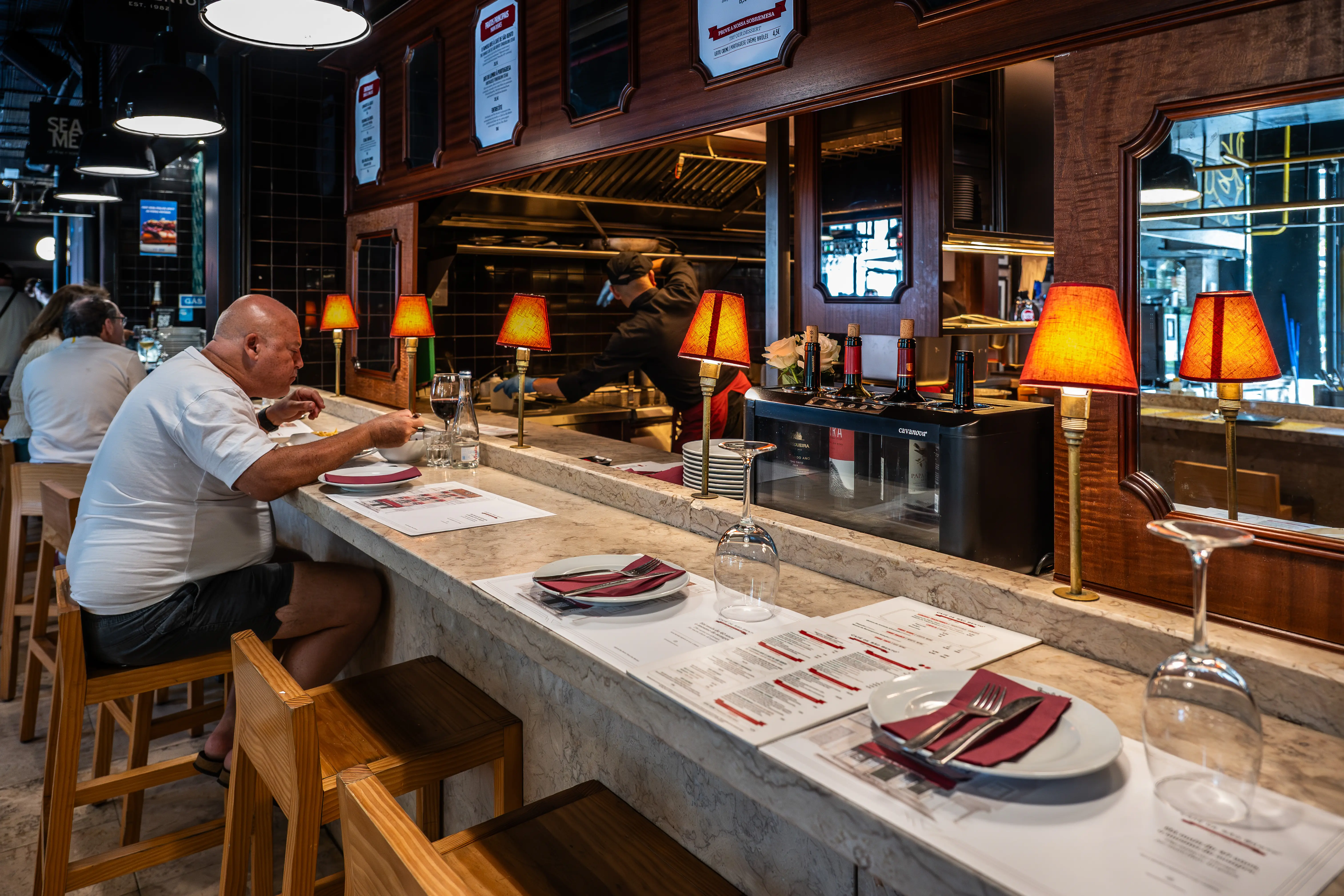 A bald man is seated at a bar, eating a meal. Other patrons are visible in the background, and a chef works behind the counter. Small lamps with red shades provide ambient lighting.