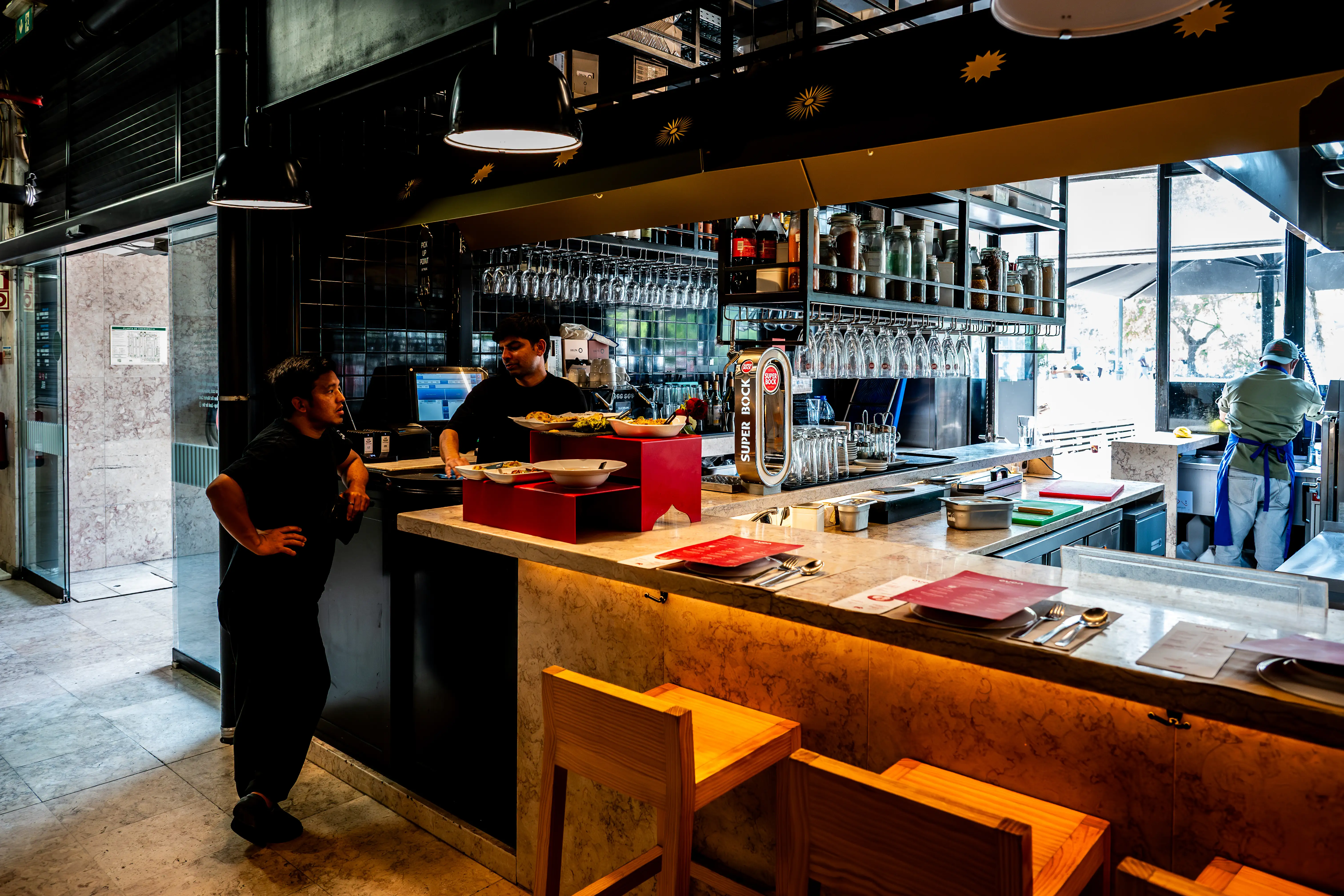  A busy restaurant bar interior with two chefs behind the counter and a person in the background working. There are also bar stools in the foreground and a large window letting in natural light.