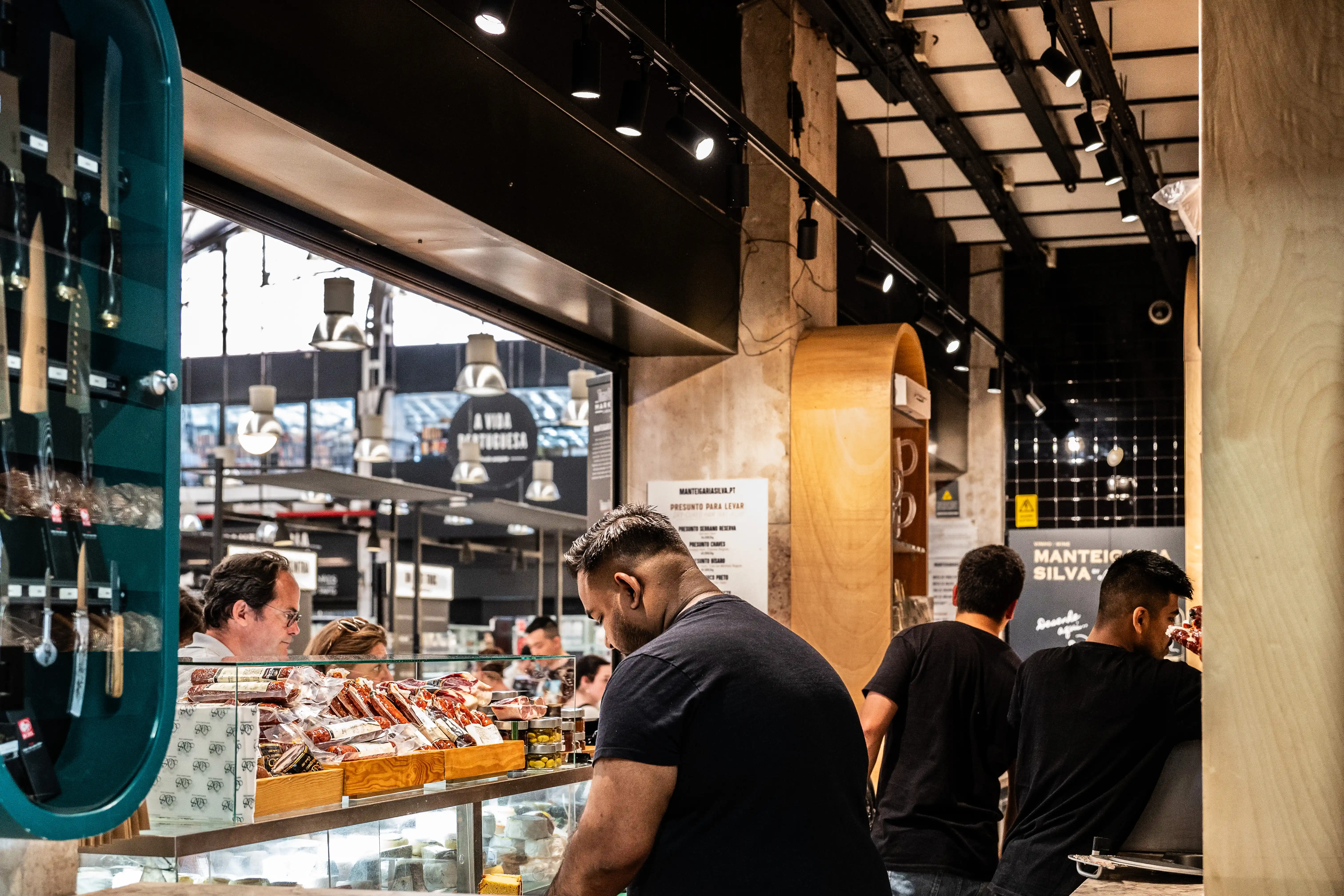 A view of a well-stocked deli counter inside a market, featuring a variety of cured meats and cheeses. Customers and staff are visible, indicating a lively atmosphere. The lighting is modern with track lights overhead.