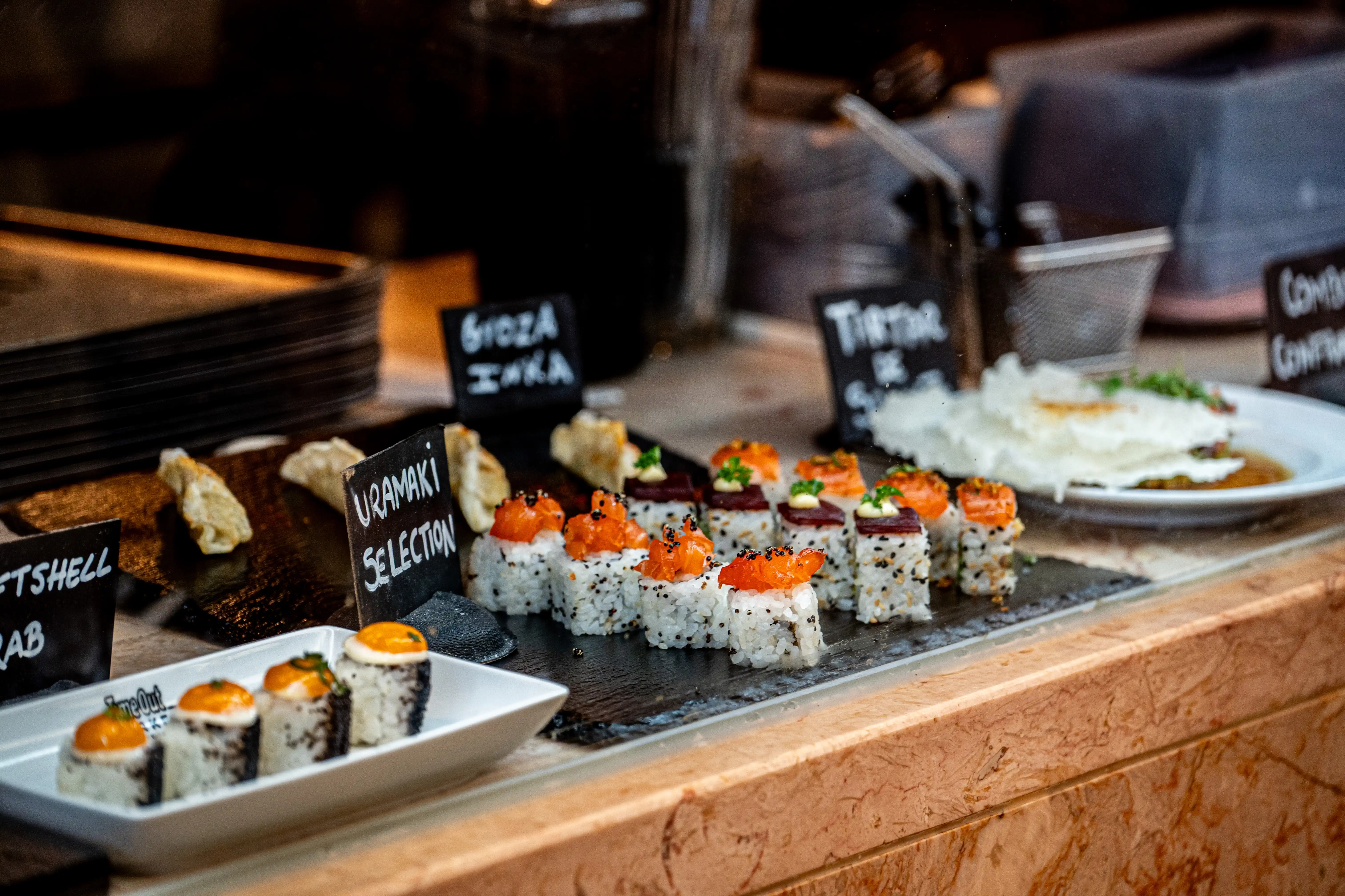 A glass display case filled with various Japanese food items, including salmon uramaki sushi rolls, sushi with orange topping, and dumplings, with handwritten labels.