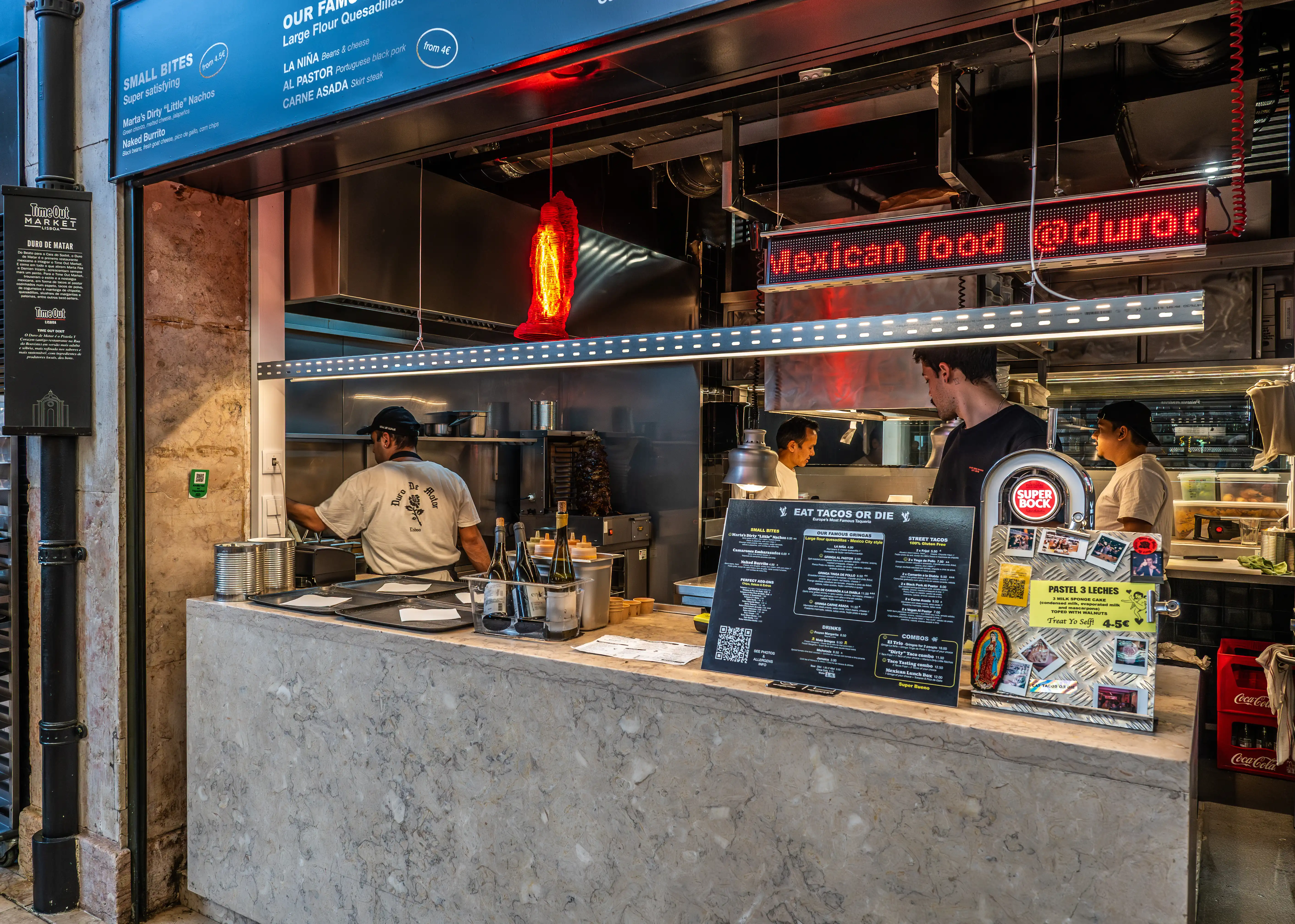 A busy Mexican food stall with staff behind the counter and menus on display. A sign above reads 