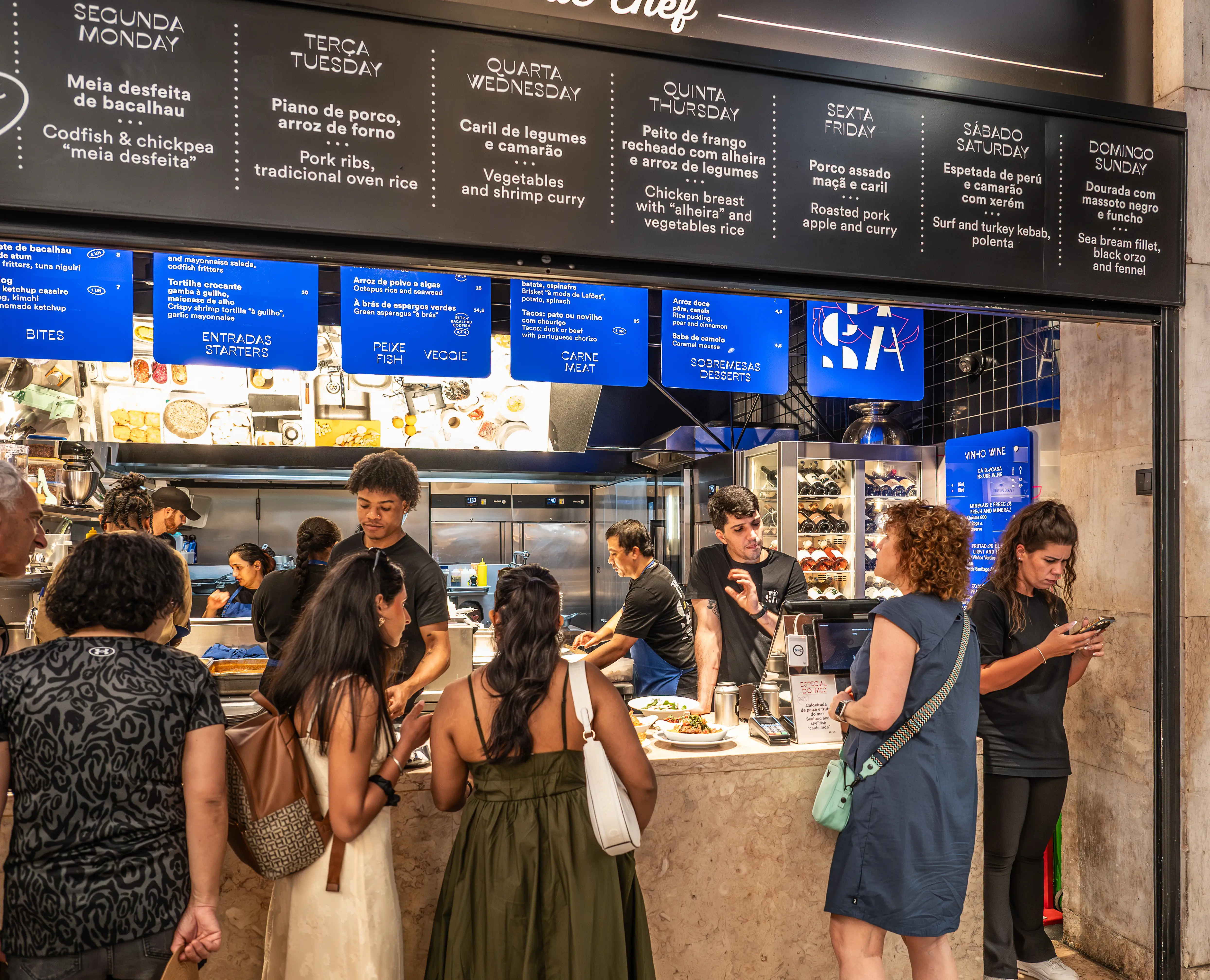 A black menu board hangs above a food counter, showcasing daily specials in Portuguese and English for Monday through Sunday. Several customers are waiting and ordering food from staff behind the counter.