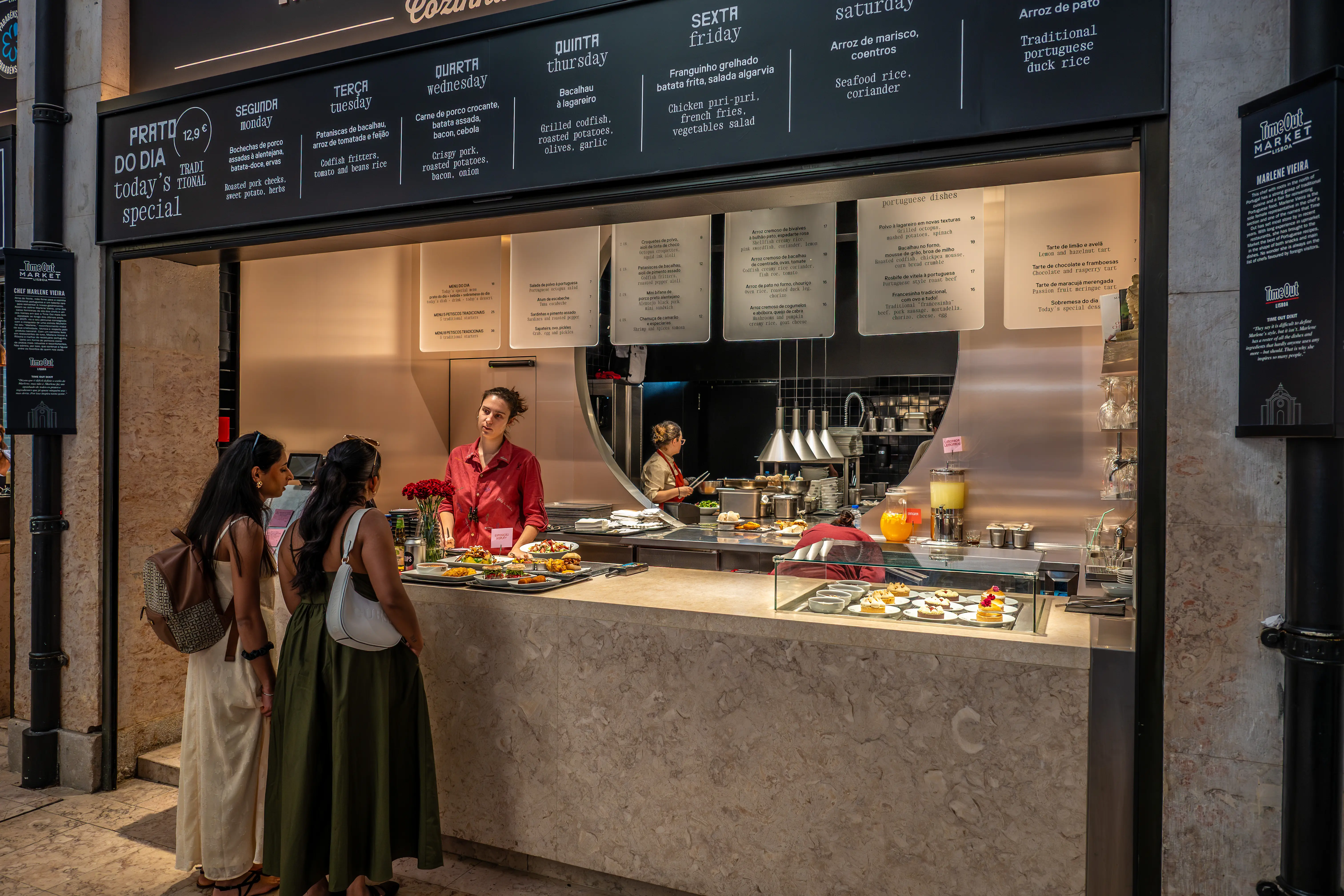 A busy food counter at Time Out Market Lisbon, with a staff member serving two customers. Menu boards display daily specials in Portuguese and English.