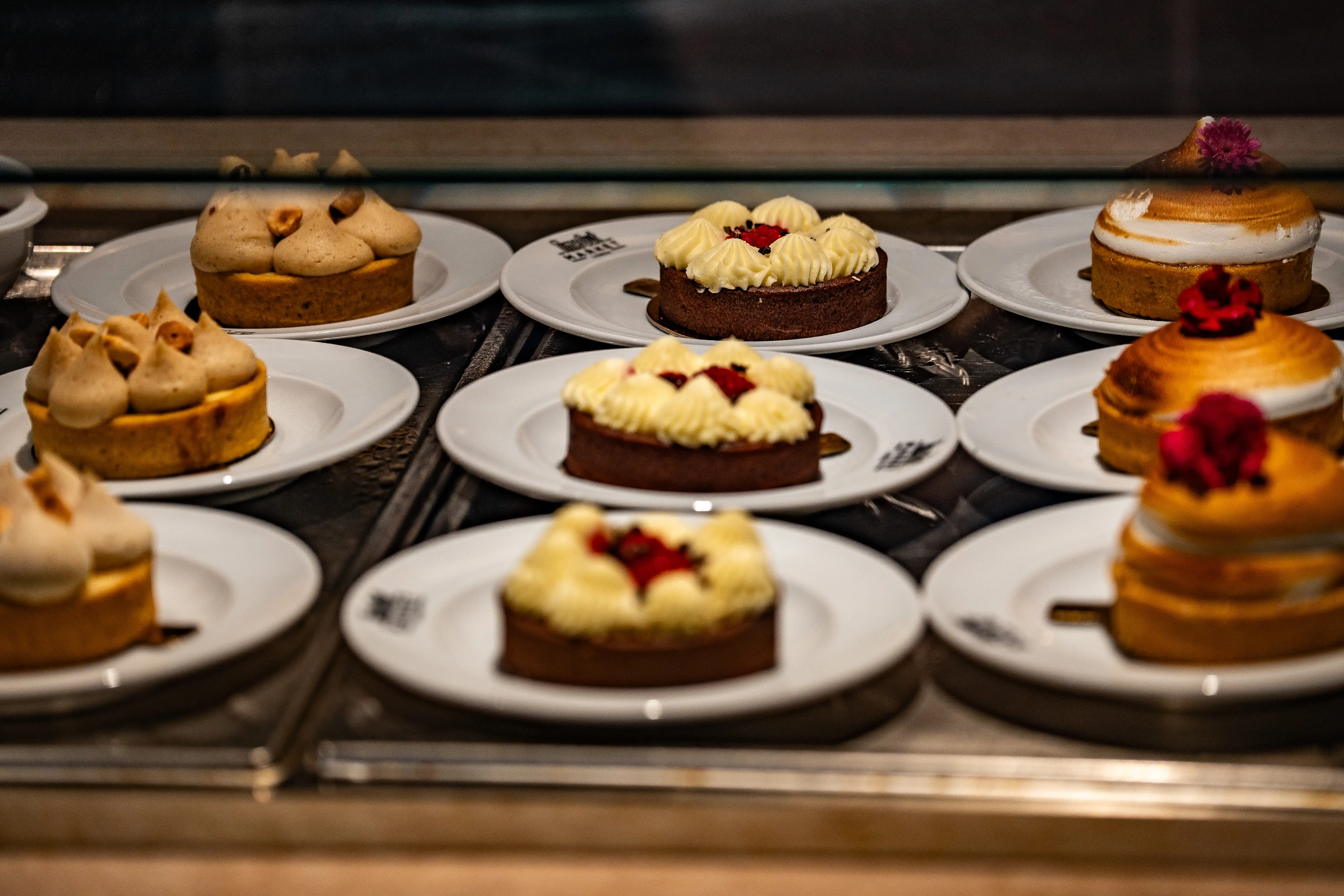 A display case filled with various small tarts and cakes, featuring different toppings like cream, nuts, berries, and meringue.