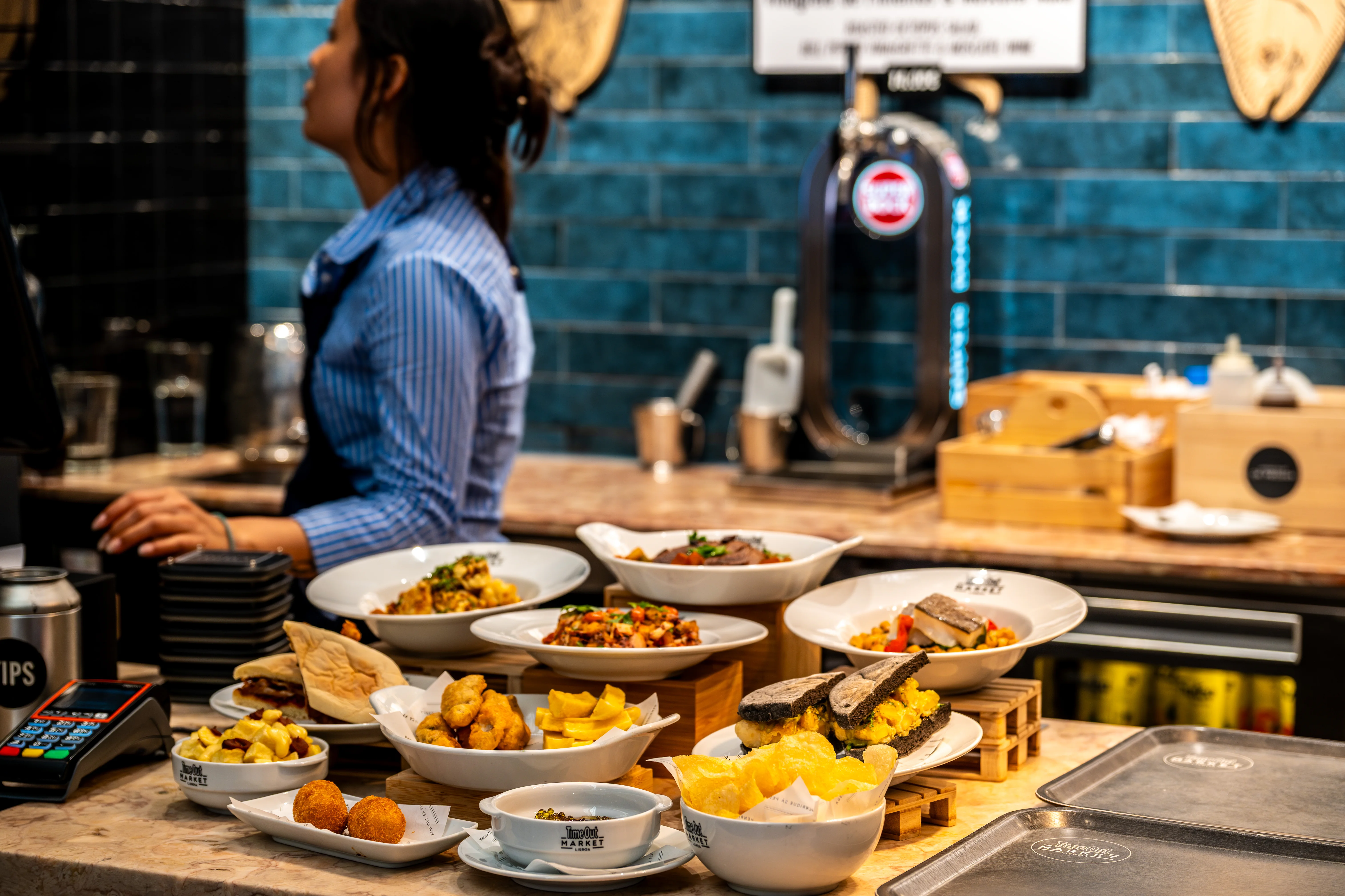 A close-up view of various small plates filled with different food items, including fried snacks, sandwiches, and salads, arranged on a counter at a food market. A woman in a striped shirt is blurred in the background.
