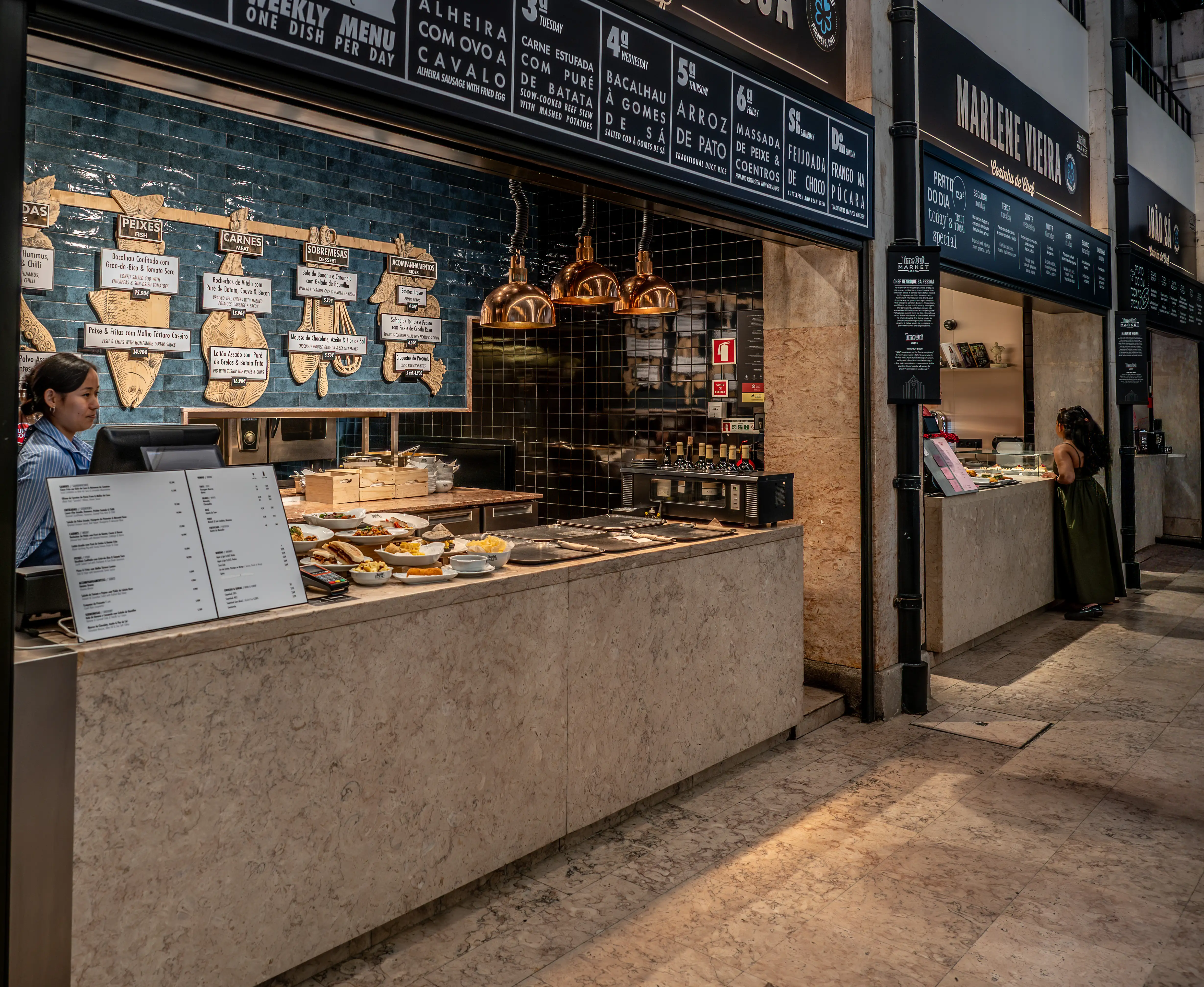 A food market stall interior with a staff member behind the counter, showcasing a variety of dishes and menu boards written in Portuguese.