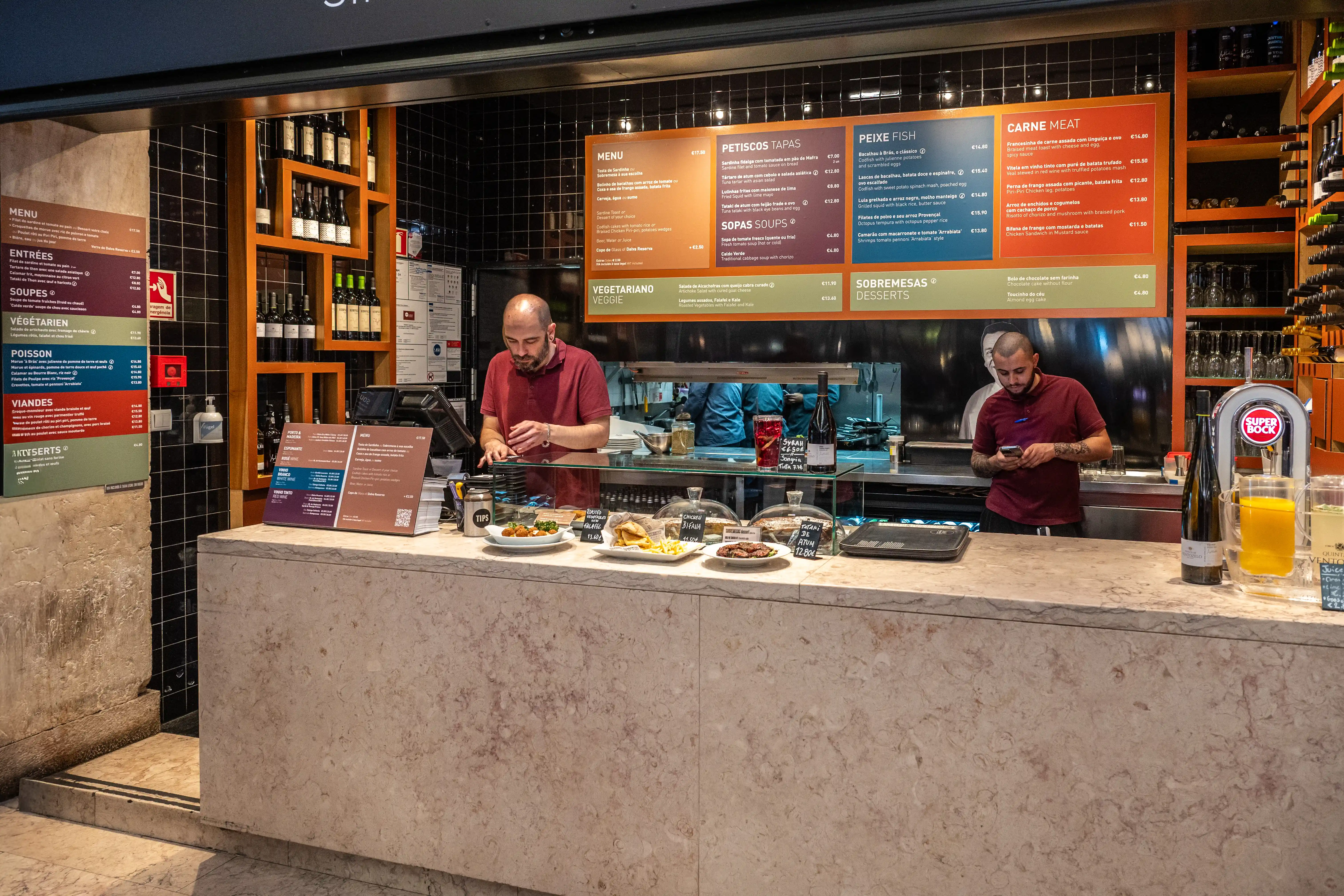A busy food counter in a Portuguese restaurant with staff members serving customers, a selection of food items displayed, and a wine rack filled with bottles in the background.