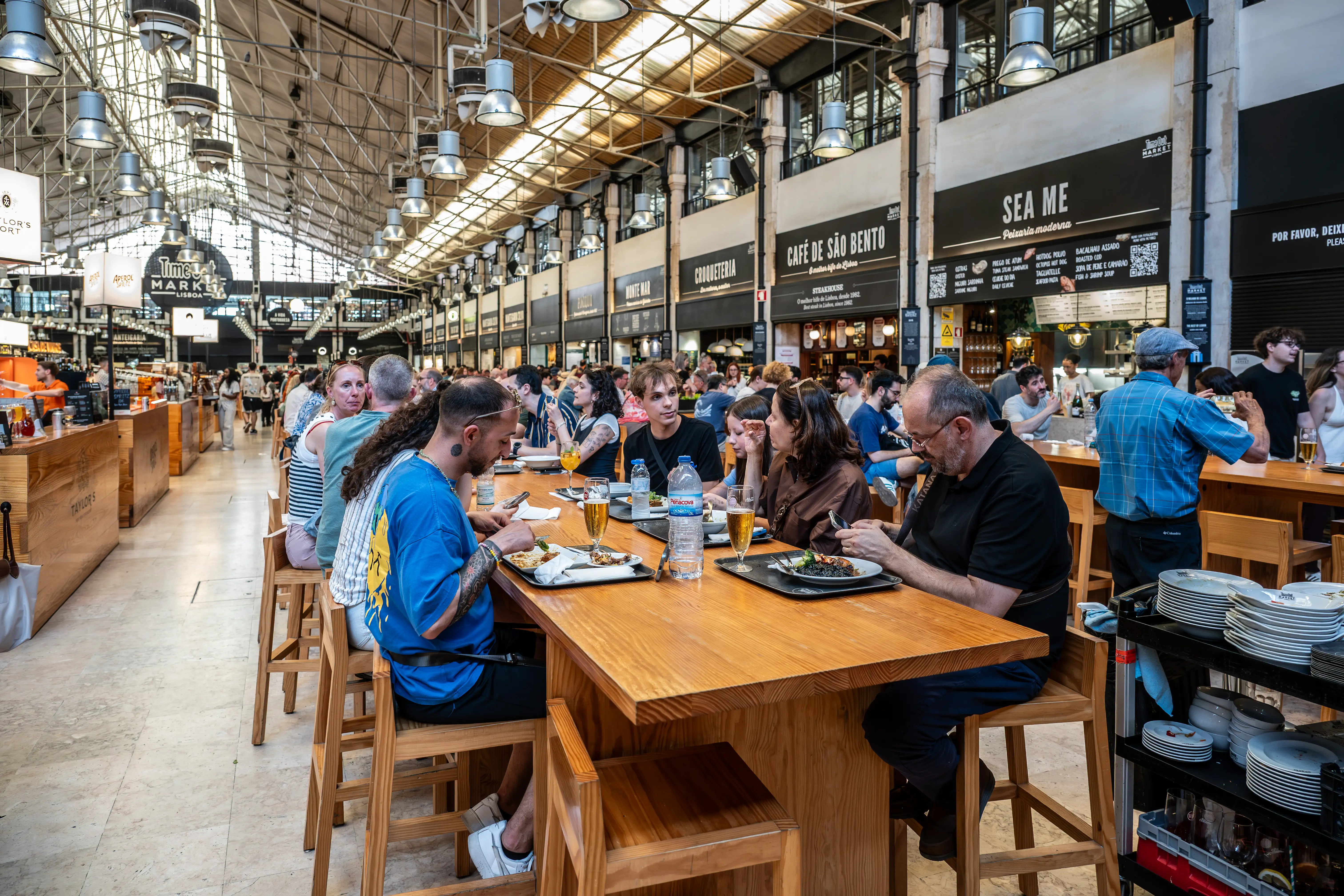 A busy food market in Lisbon with people dining at long wooden tables. Several food stalls with menus and signage are visible in the background.
