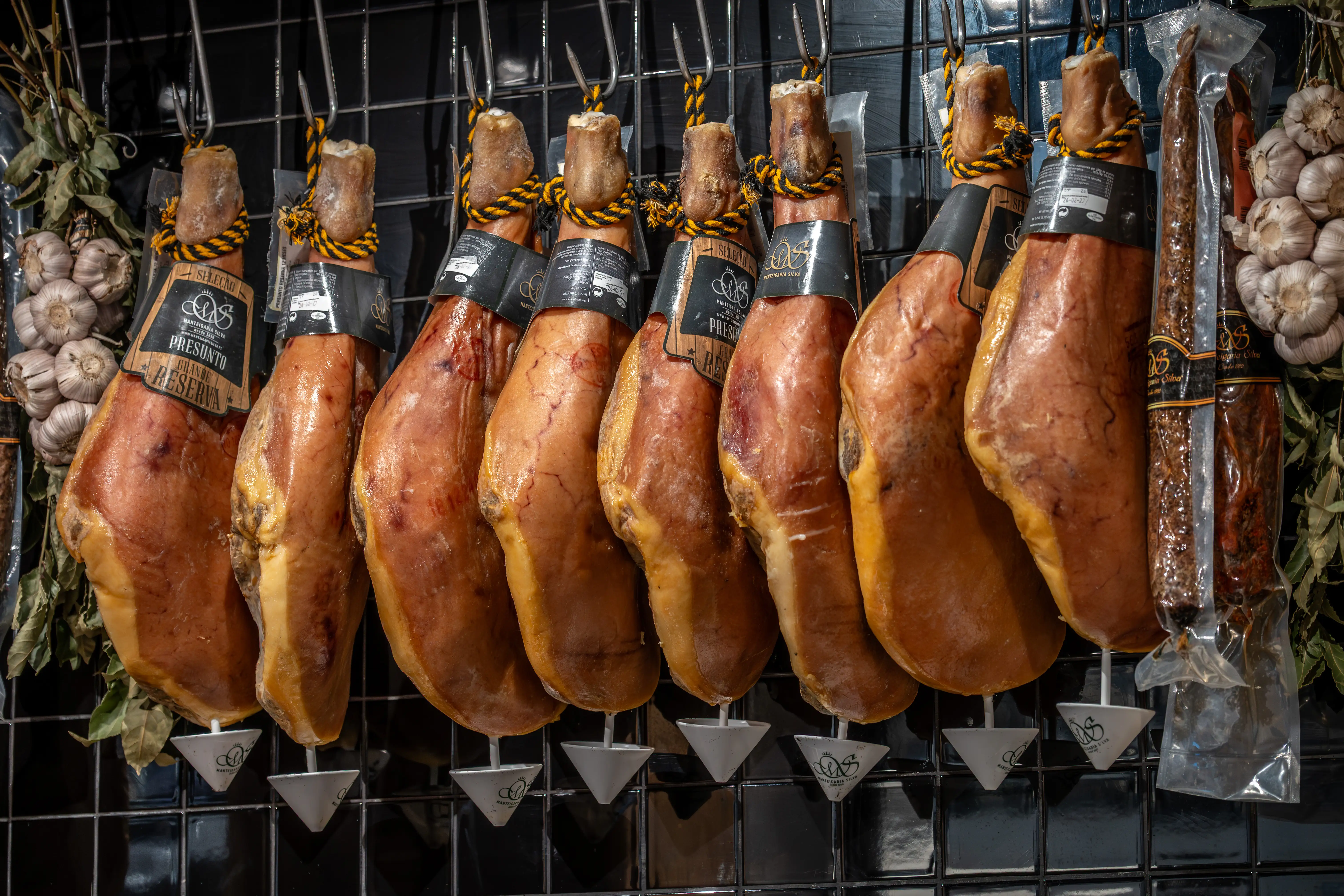 A selection of whole Iberian hams, each with a branded label and tied with rope, hang in a row, with cured sausages and garlic bunches visible.