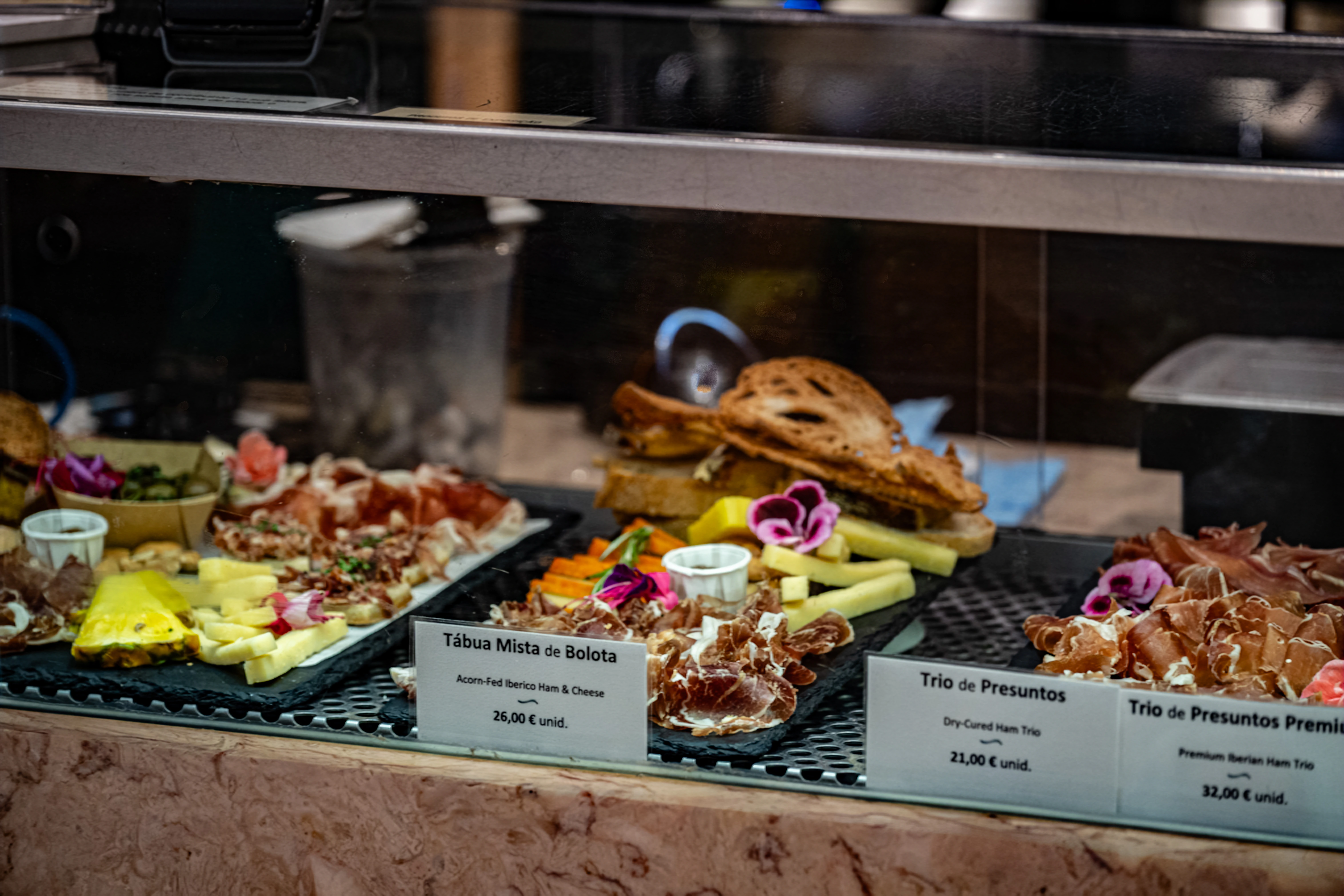 A chilled display counter filled with appetising platters of thinly sliced Iberian ham and various cheeses, garnished with edible flowers and accompanied by price labels.