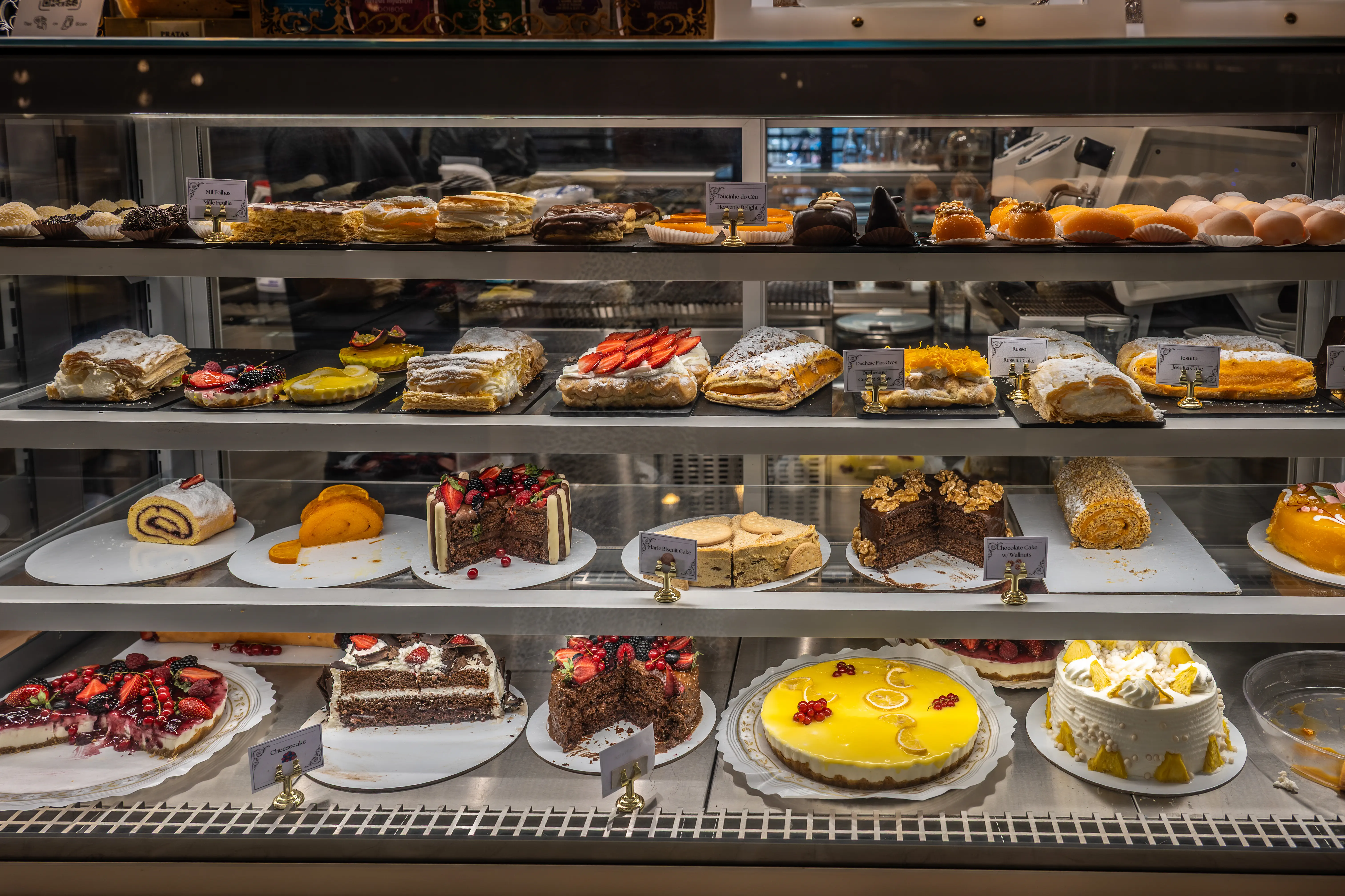 : A refrigerated display cabinet filled with various pastries and cakes, including fruit tarts, layered cakes, and roulades, presented on multiple shelves.
