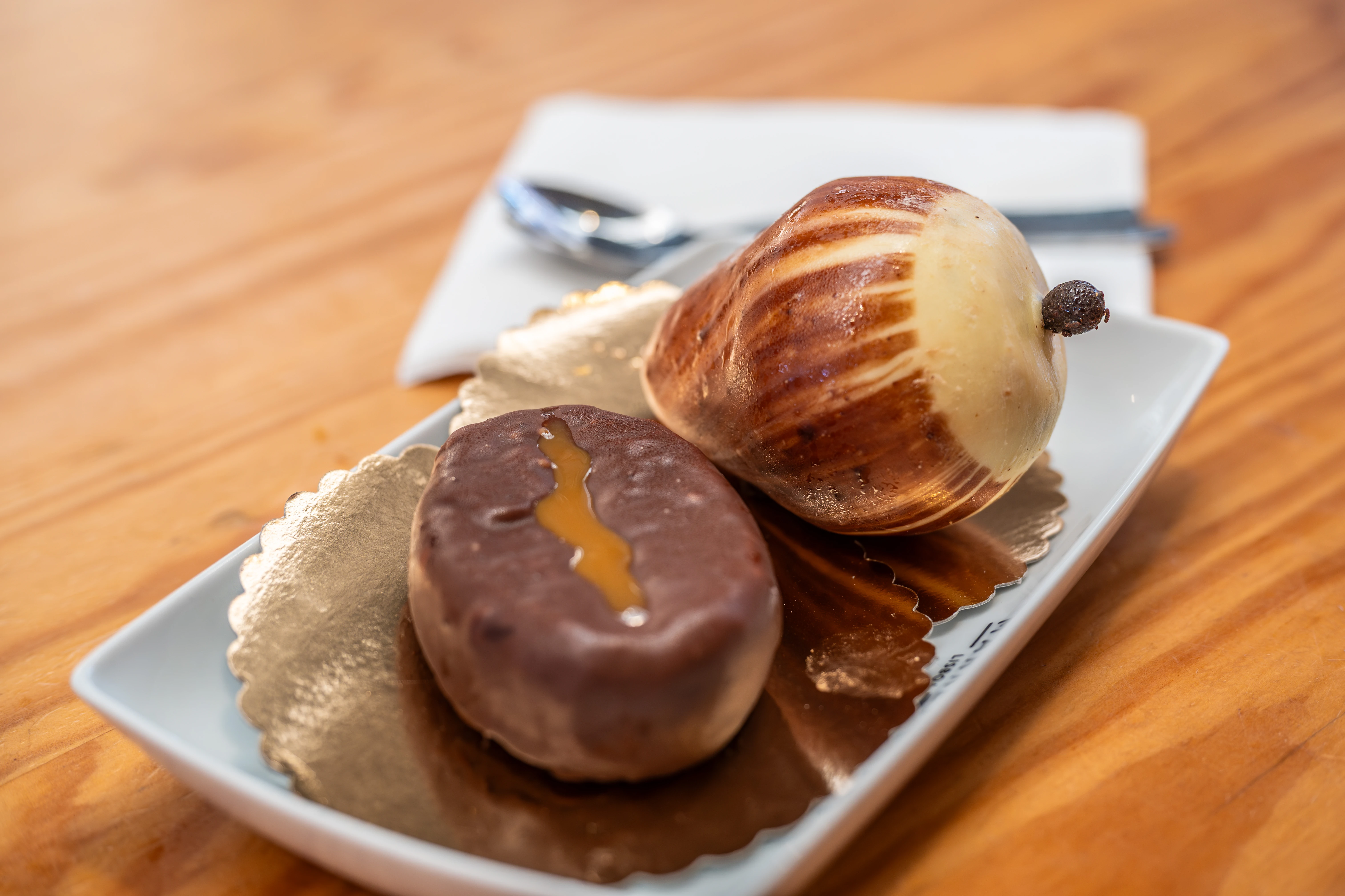 Two artistic pastries, a chocolate oval with caramel and a striped pear-shaped marzipan, on a white plate with gold liner.
