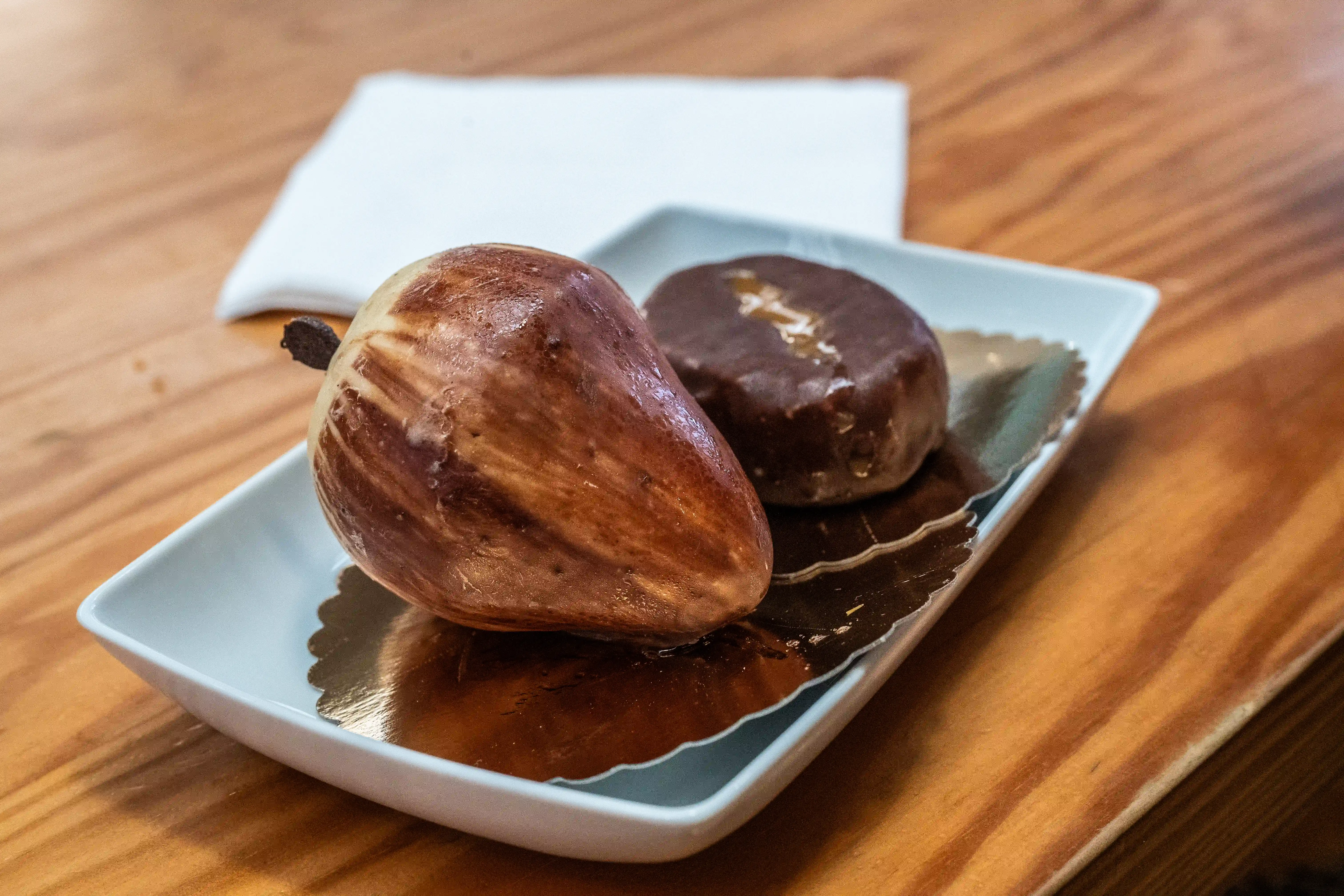  Two decorative pastries, one pear-shaped with marbled icing and another round chocolate-covered pastry, served on a white dish with gold trim.