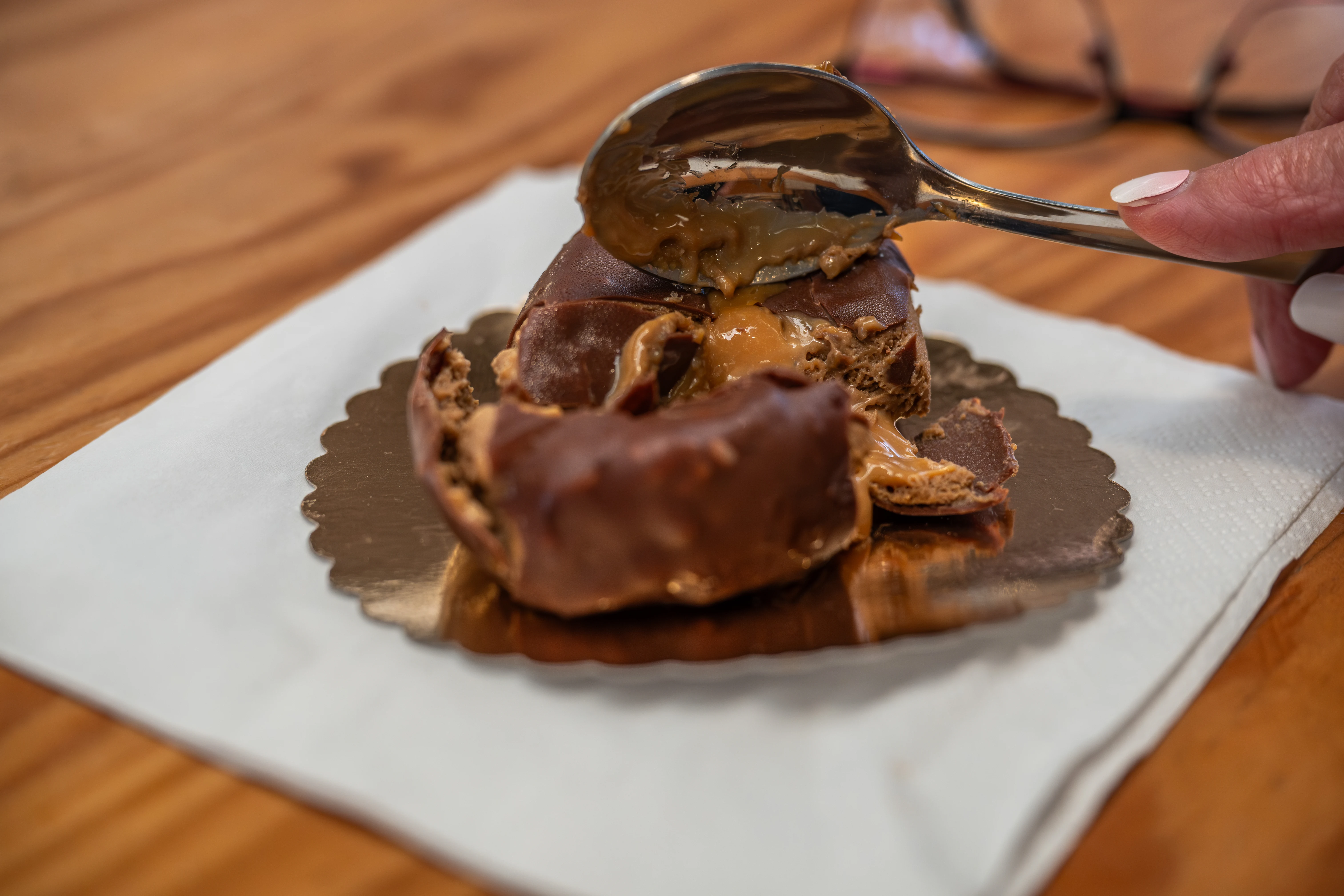 A person's hand with a manicured fingernail uses a spoon to scoop into a chocolate pastry that has a gooey caramel centre.