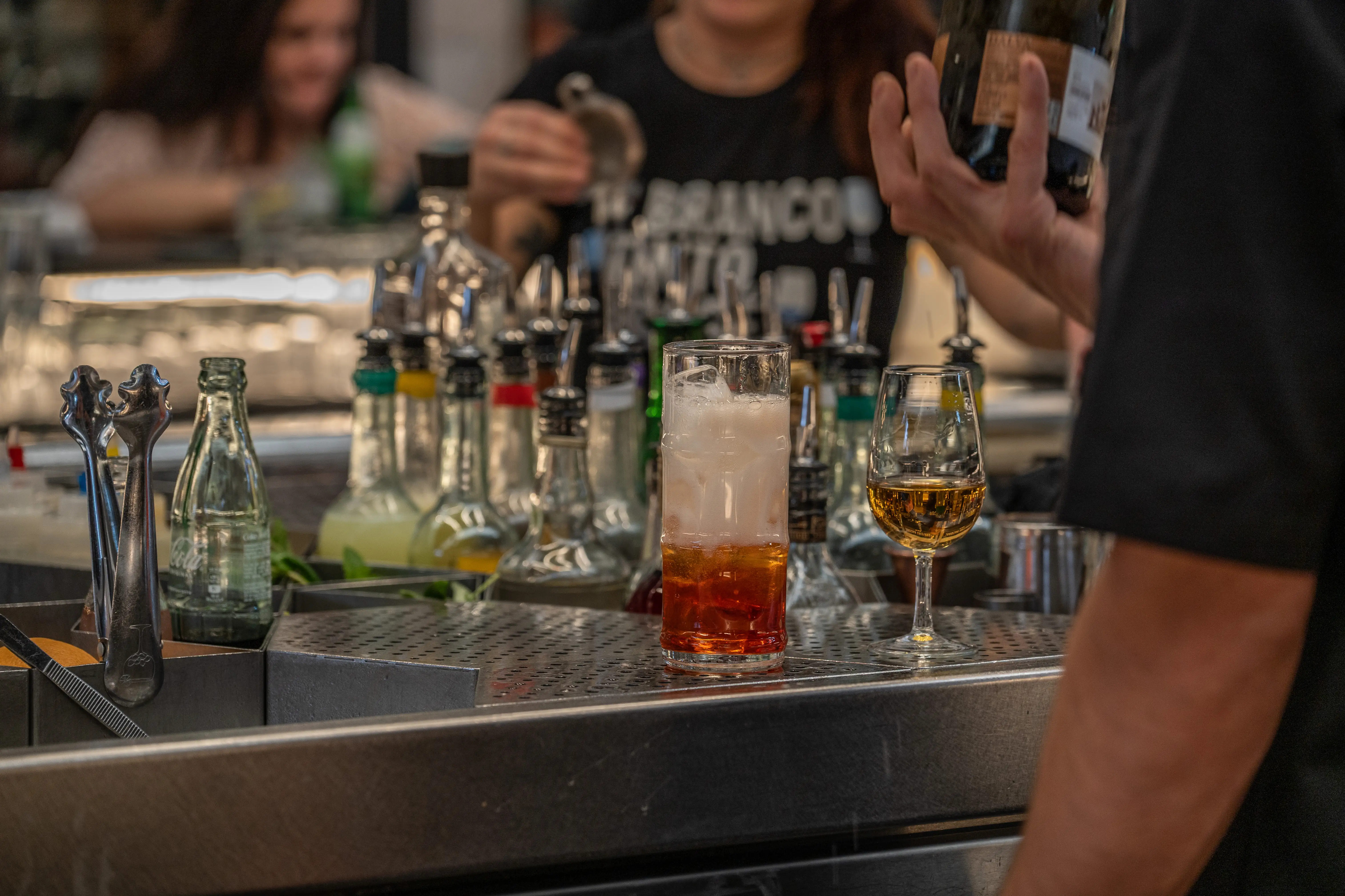 A bartender prepares drinks behind a bar, with a layered cocktail, a tasting glass of amber spirit, and numerous spirit bottles visible.