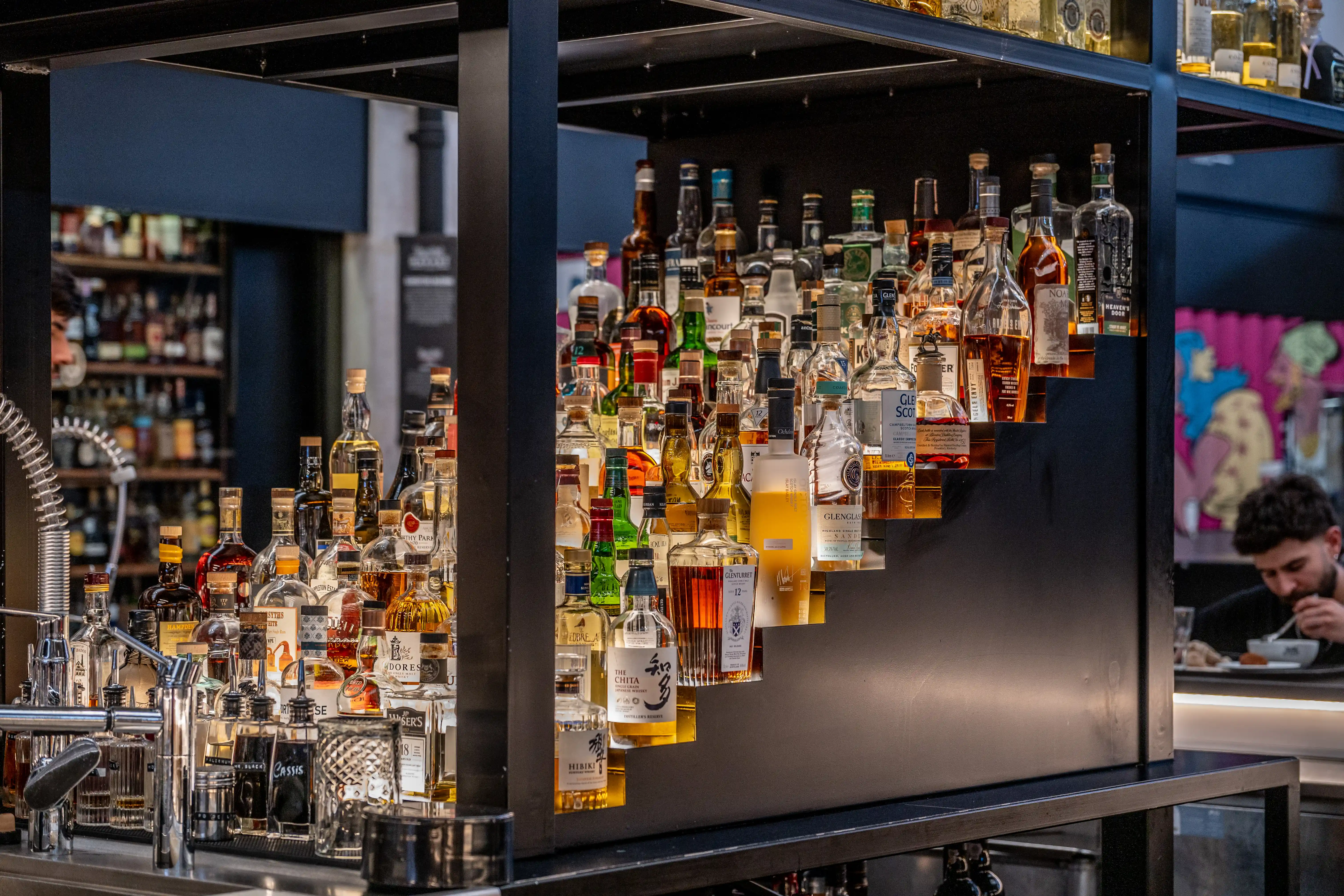 A tiered shelf display overflowing with bottles of spirits, including various whiskies and gins, in a bar. A man is eating in the background.