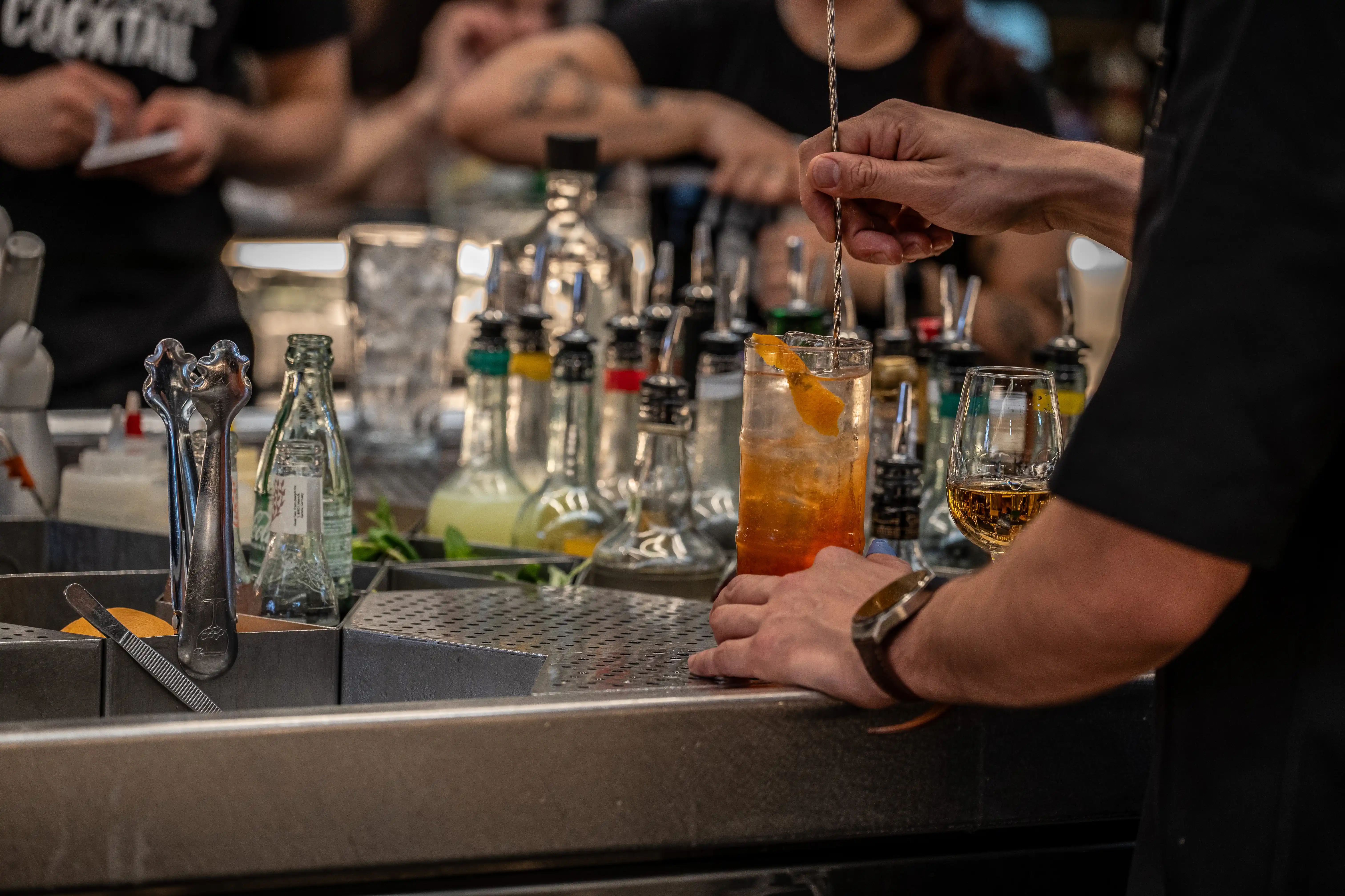  A blurred image of a bar counter with bottles of colourful mixers, a glass of ice, and a bartender in the background.