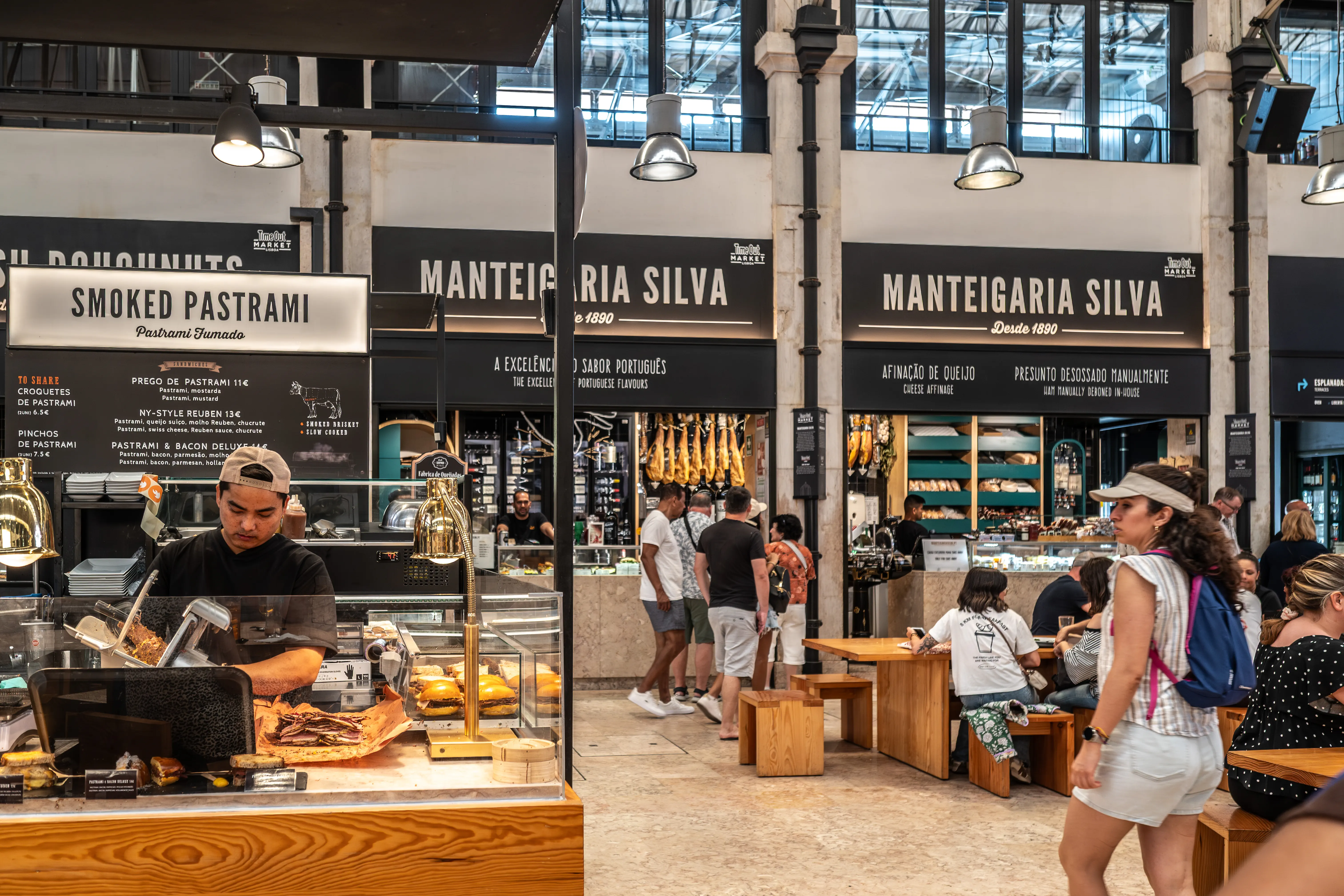 A busy food market with people browsing stalls. A vendor prepares pastrami at a counter, with sandwiches and burgers on display. Another stall features hanging cured meats and cheeses. Customers are seated at wooden tables in the background.