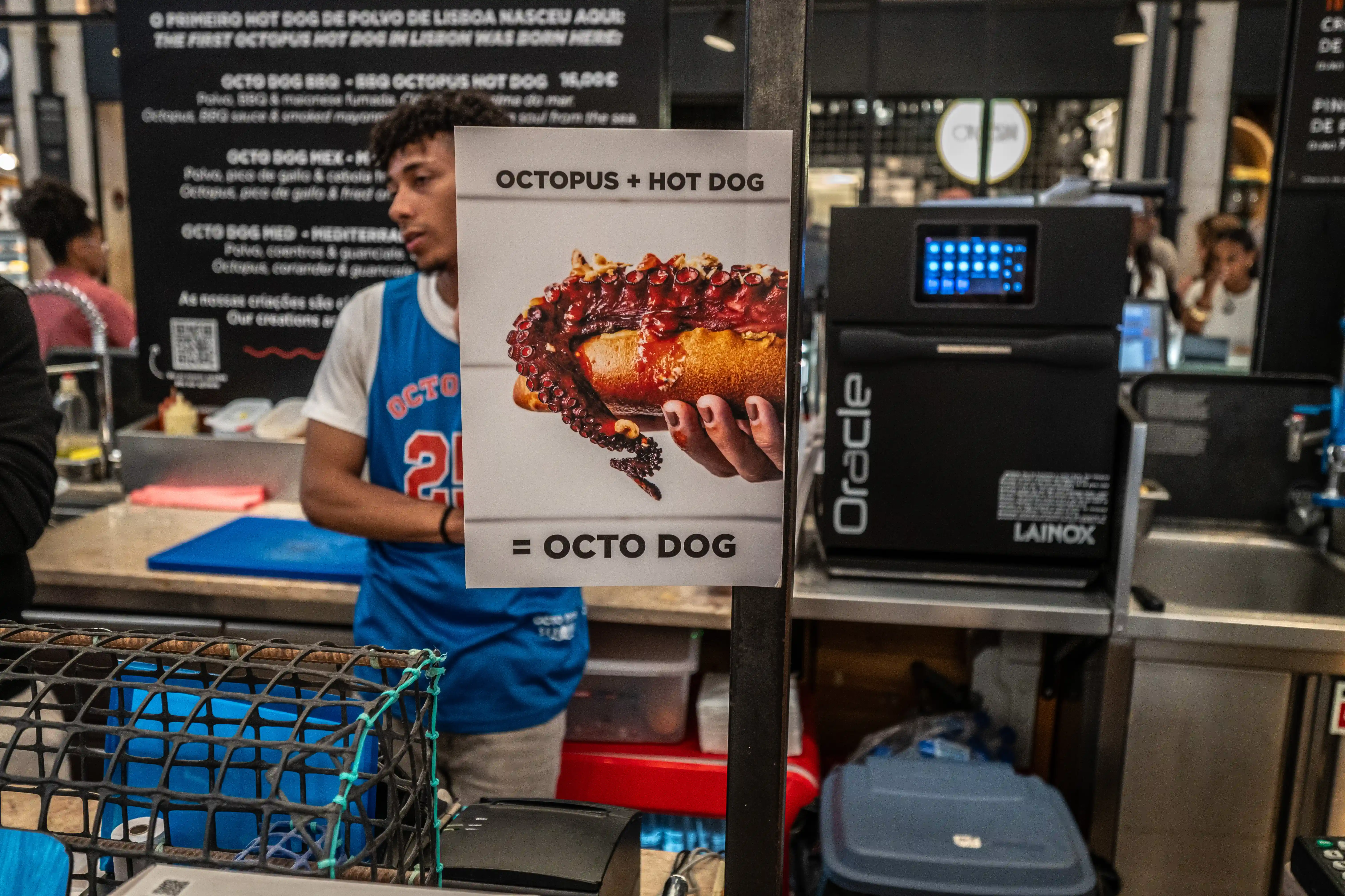A close-up of a sign advertising an Octo Dog, which is a hot dog topped with octopus tentacles, held by a hand. A young man wearing a blue OCTO vest stands behind the sign.