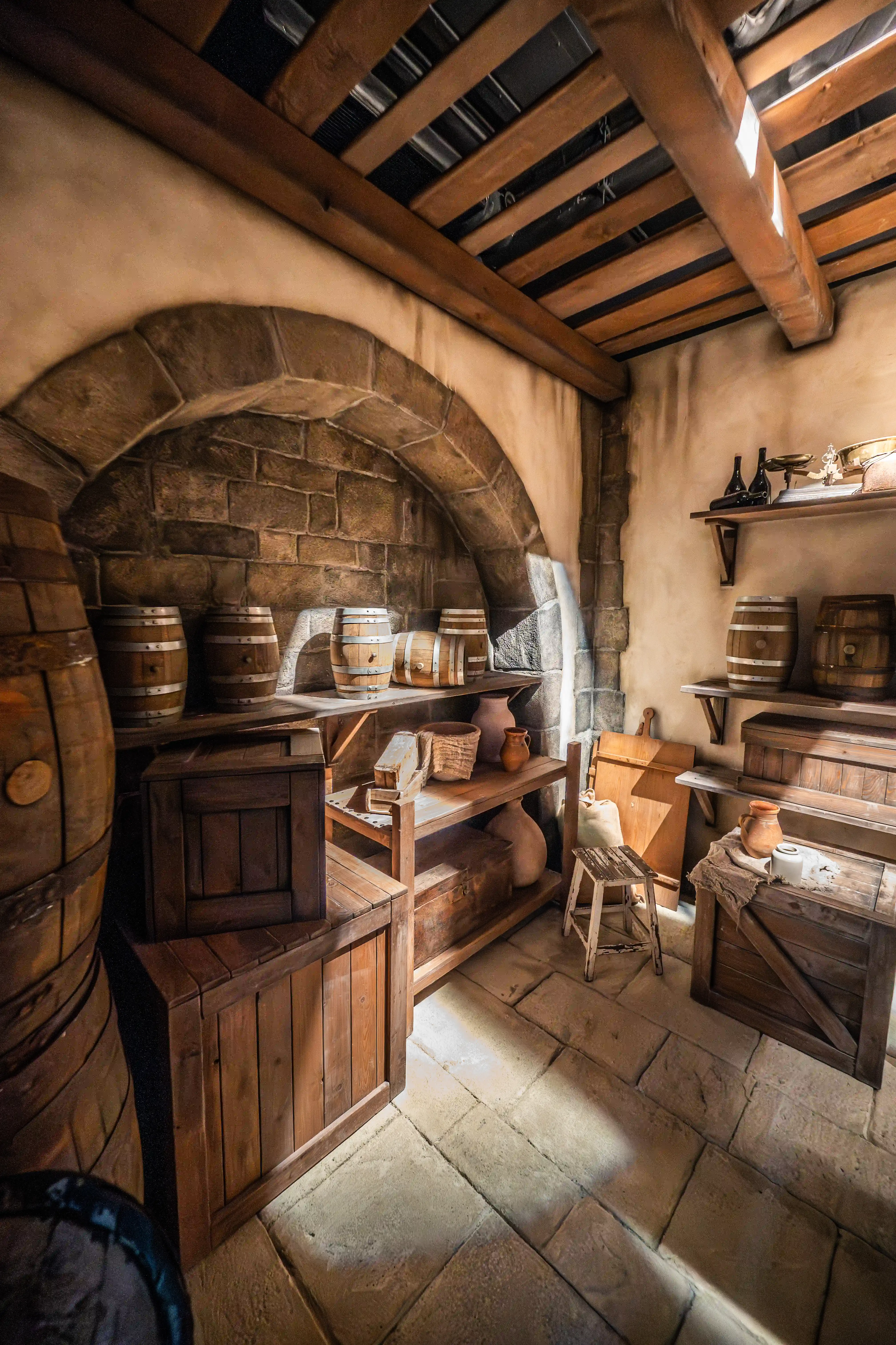 Interior of a rustic cellar with stone walls and arched entryway, filled with wooden barrels, crates, and shelves. Beams of sunlight streak across the floor and illuminate parts of the room.