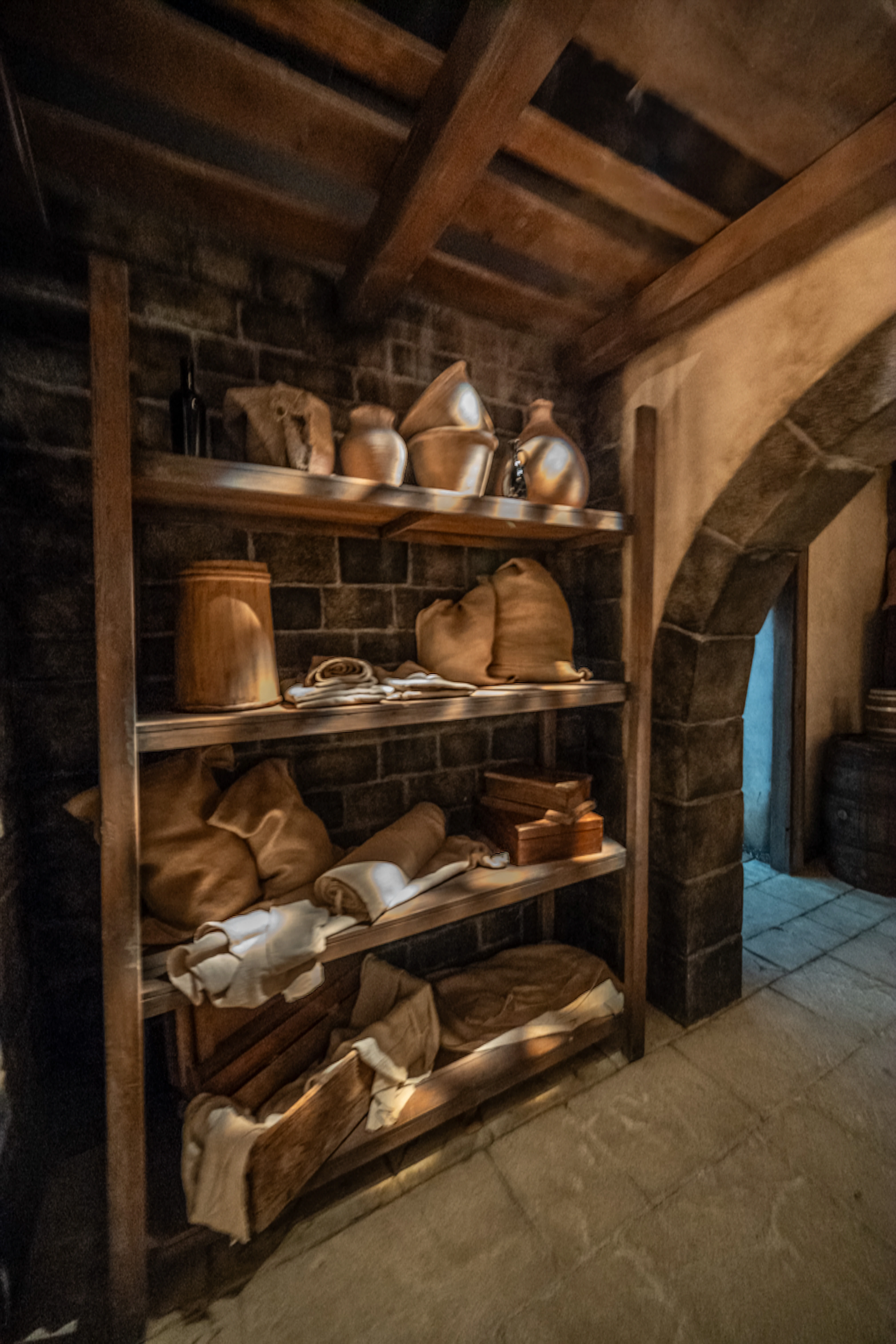 Wooden shelves in a rustic room are filled with sacks, pottery, and boxes. Sunlight streams down, creating strong shadows and highlights. An arched stone doorway is visible on the right.