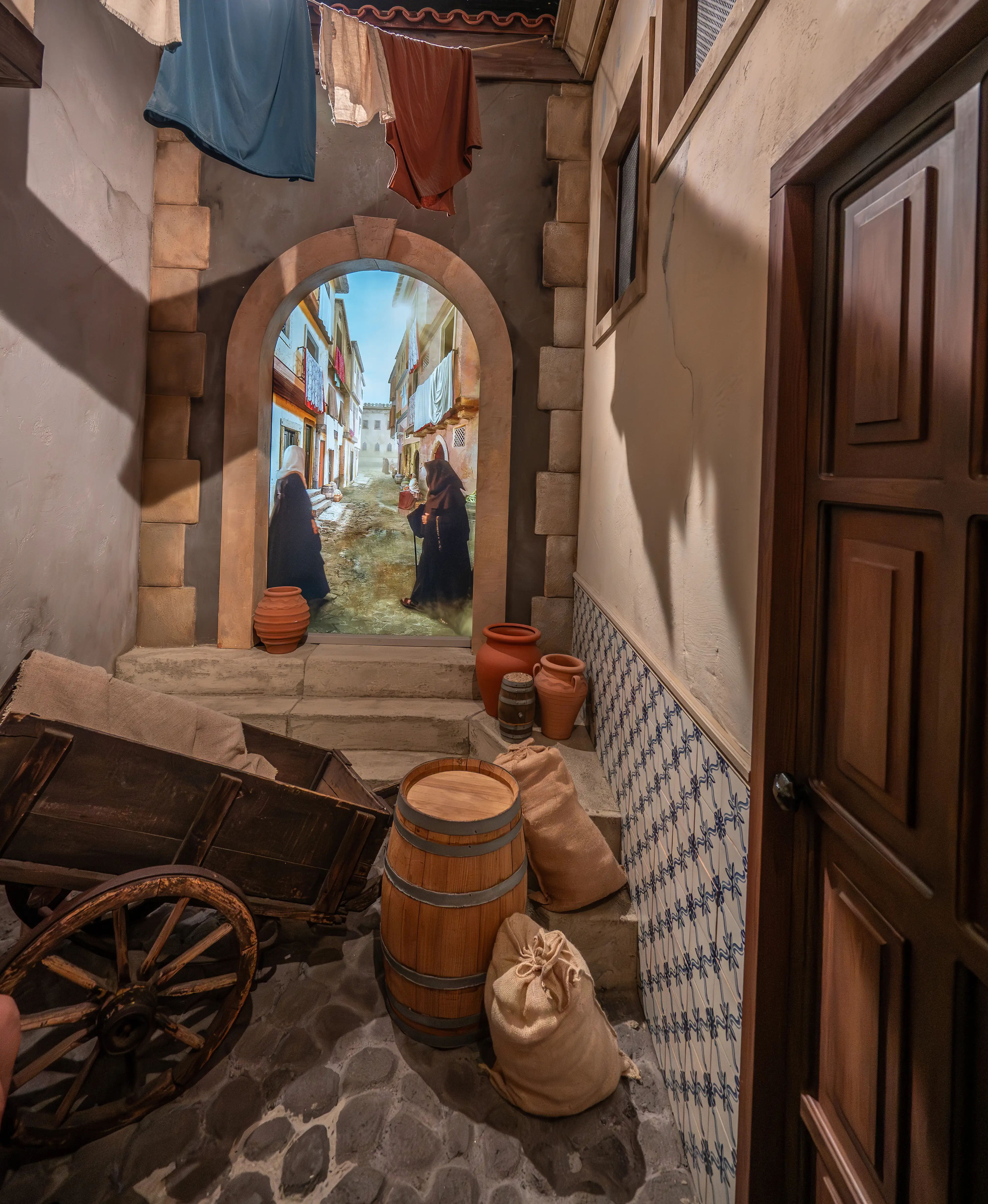 A staged scene of a medieval European alley, featuring laundry drying on a line, two robed figures entering an archway, and various objects like pottery and a cart in the foreground.