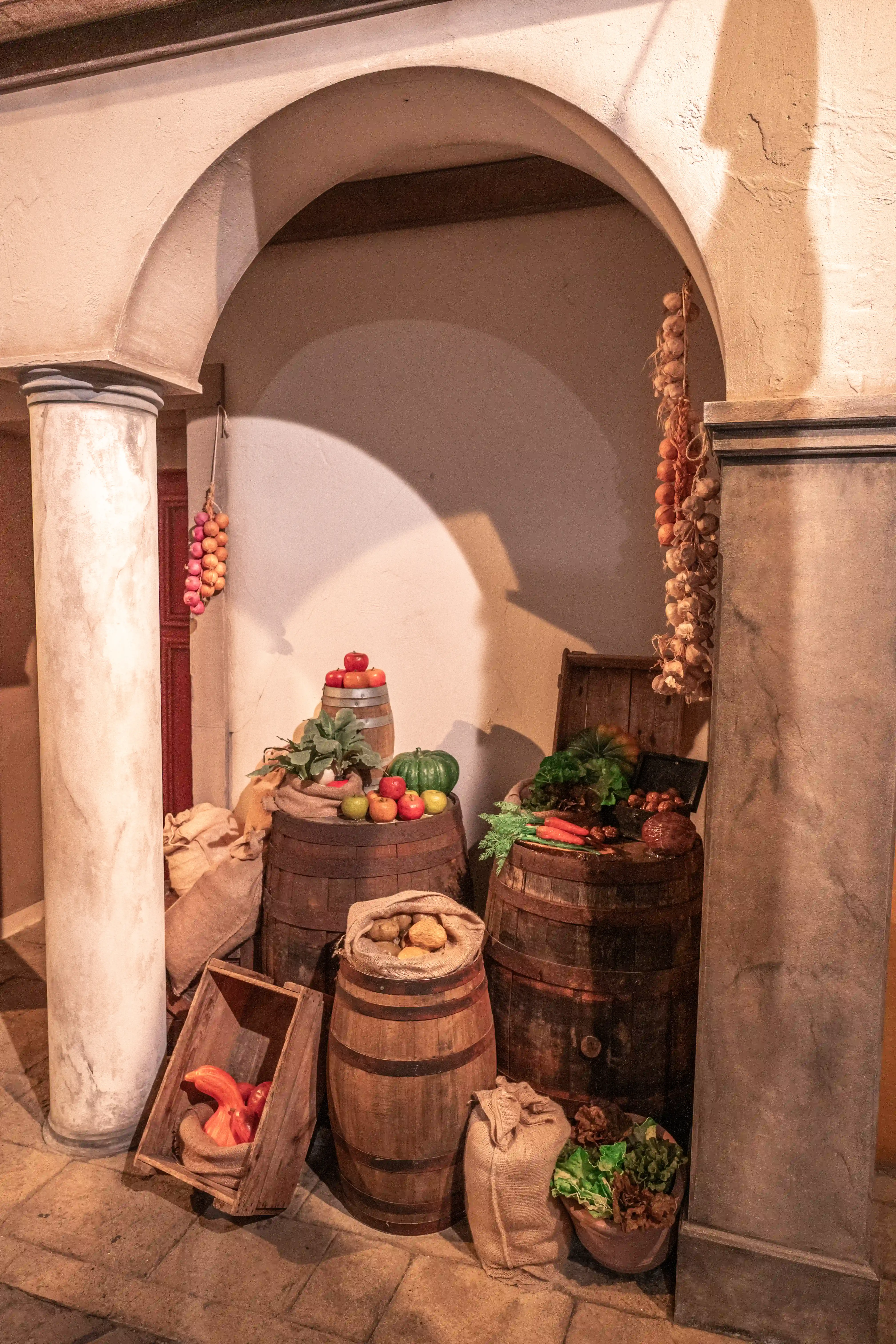 An arched entryway with a column on the left frames a rustic display of produce. Two wooden barrels are central, one topped with apples and a green pumpkin, the other with carrots and leafy greens peeking from a wooden chest. Sacks of produce and a wooden crate containing red vegetables complete the scene, with strings of onions and garlic hanging from the ceiling