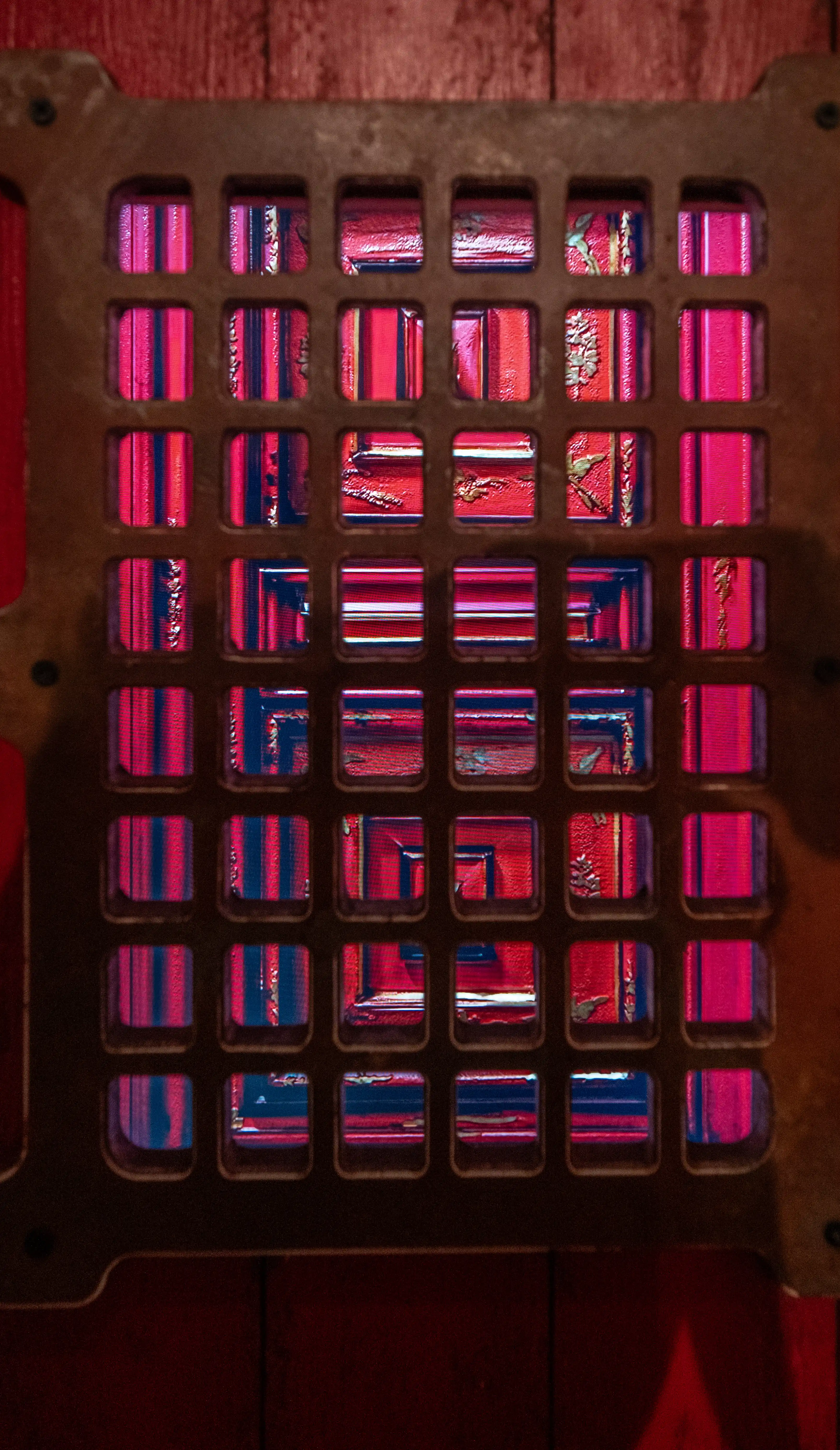  A close-up of a dark, square metal grate with a grid of square openings. Through the openings, abstract patterns of bright pink and red vertical stripes and textures are visible.