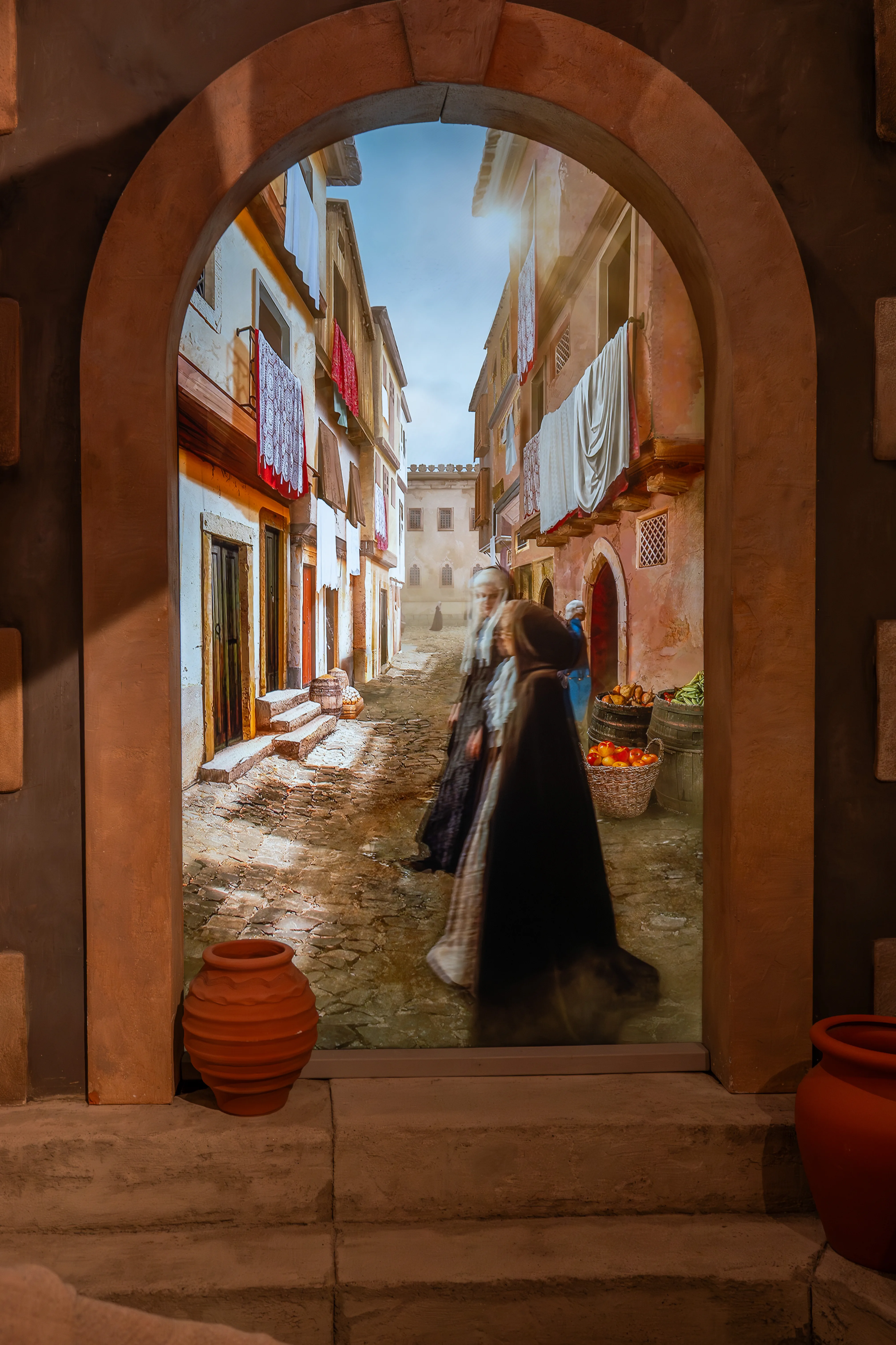 View through an arched stone doorway of a narrow, sunlit cobblestone street in an old town, with buildings on either side featuring laundry drying from windows. Two figures in dark, long clothing are walking away from the viewer, with baskets of fruit and vegetables visible.