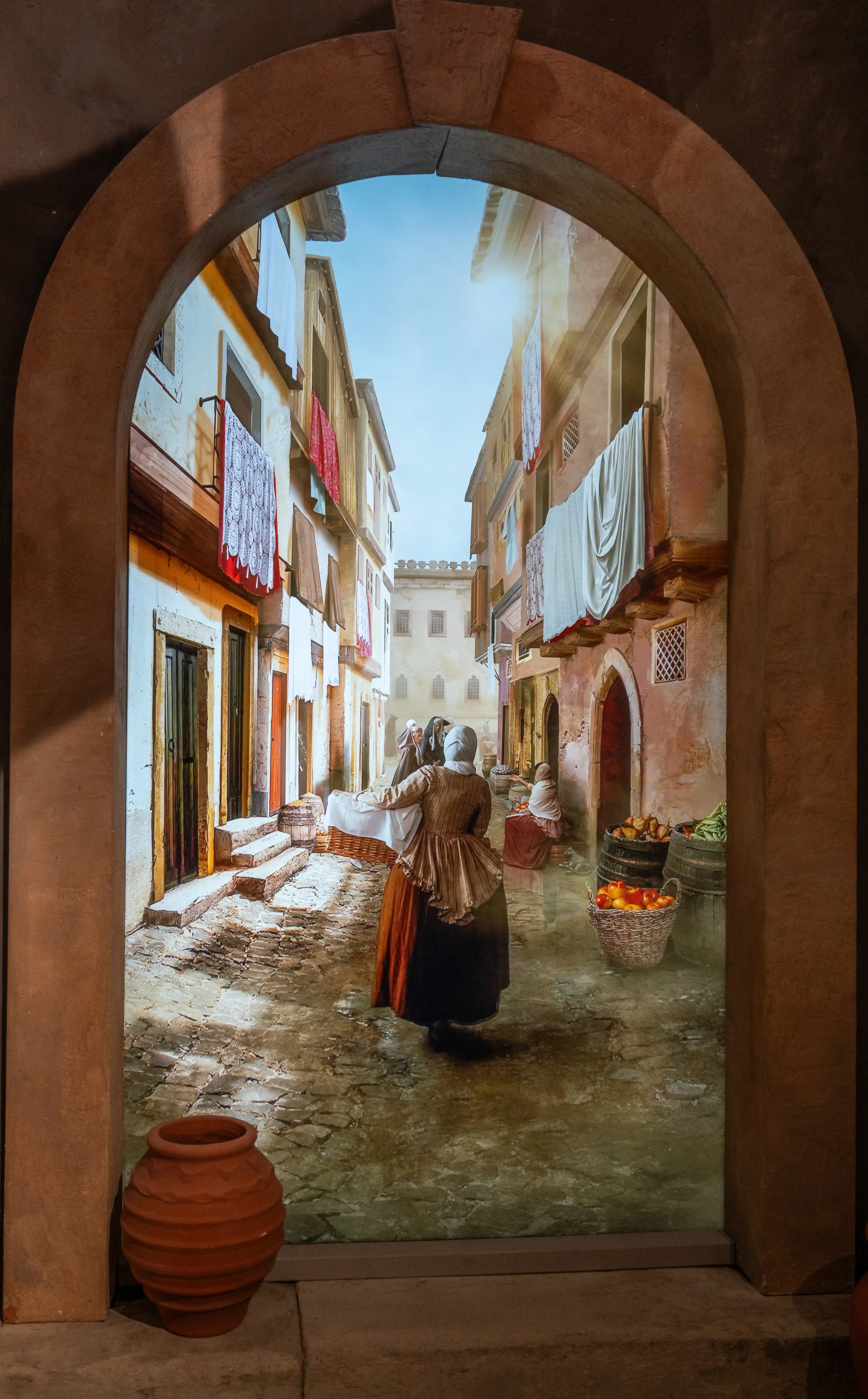 View through an arched stone doorway of a narrow, sunlit cobblestone street in an old town, with buildings on either side featuring laundry drying from windows. Two figures in dark, long clothing are walking away from the viewer, with baskets of fruit and vegetables visible.