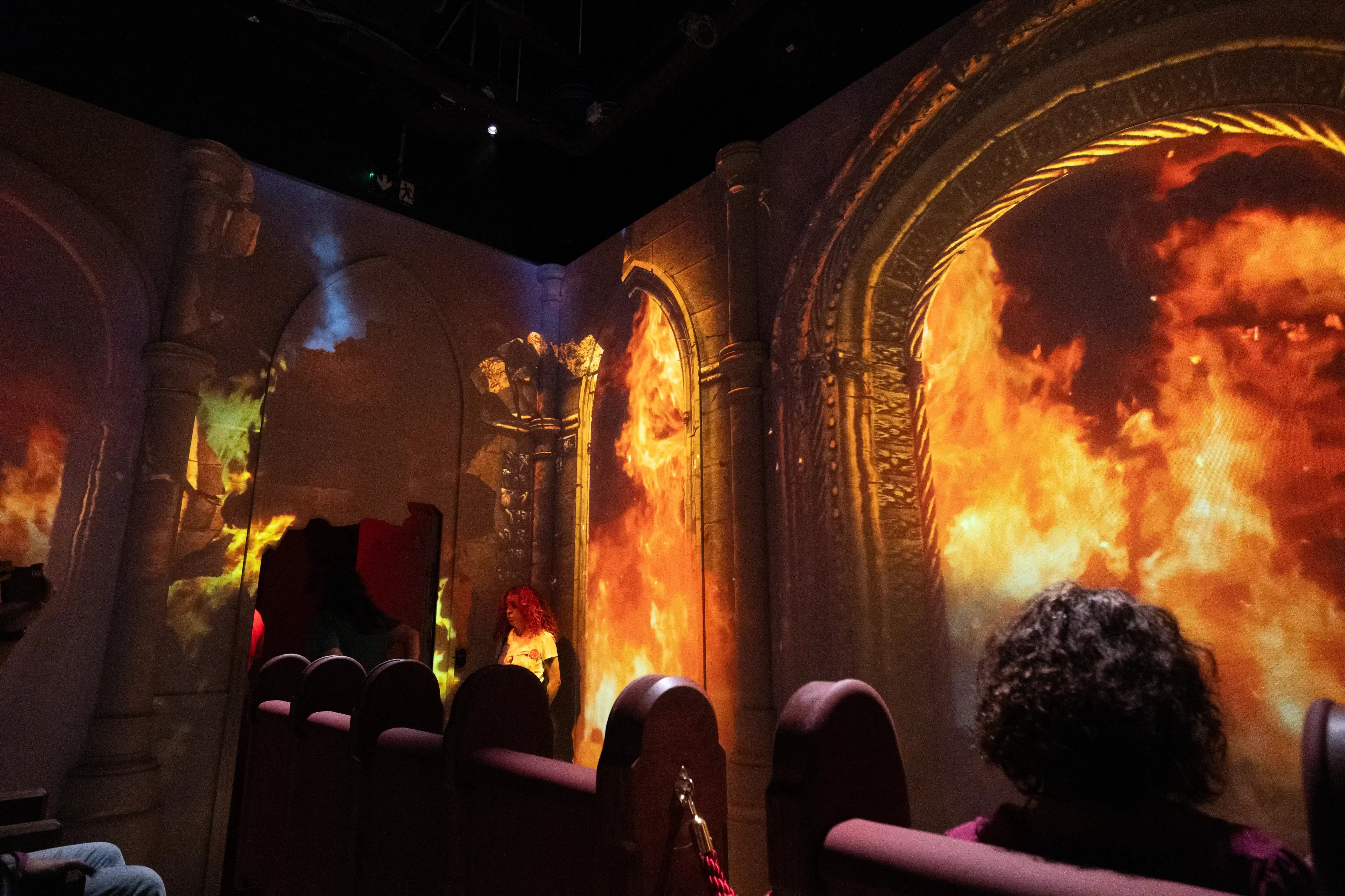 A dimly lit room with theatre-style seating faces a large projection on the wall. The projection depicts a grand stone archway filled with intense, fiery flames that appear to be engulfing the space. Architectural columns and details are visible, enhanced by the dynamic lighting of the fire effect. Several people are seated, observing the spectacle, with one individual in the middle ground standing and looking at the projection.