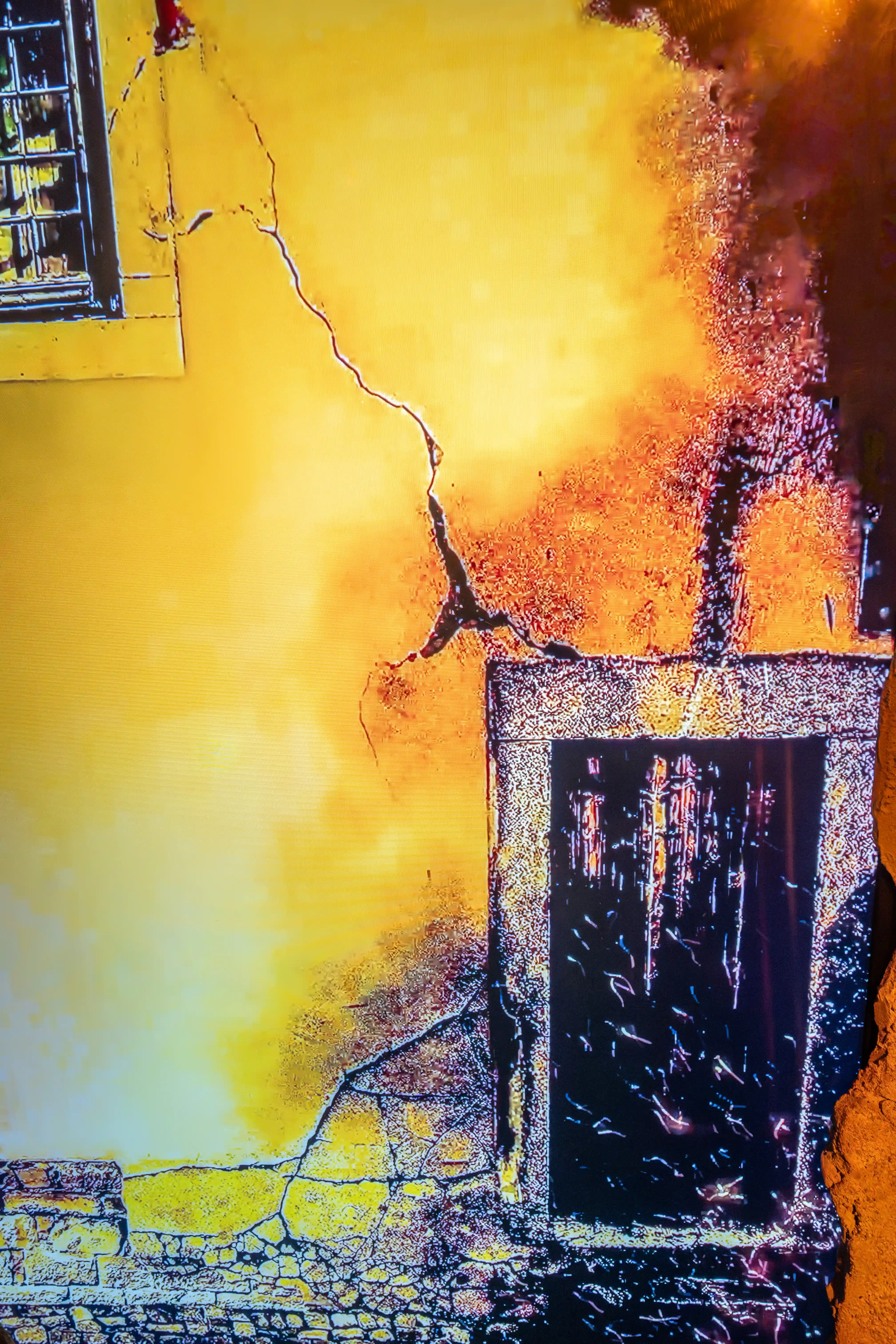 A textured image of a dilapidated building wall, showing a large crack, a barred window, and a wooden door, with areas of yellow, orange, and black discolouration.