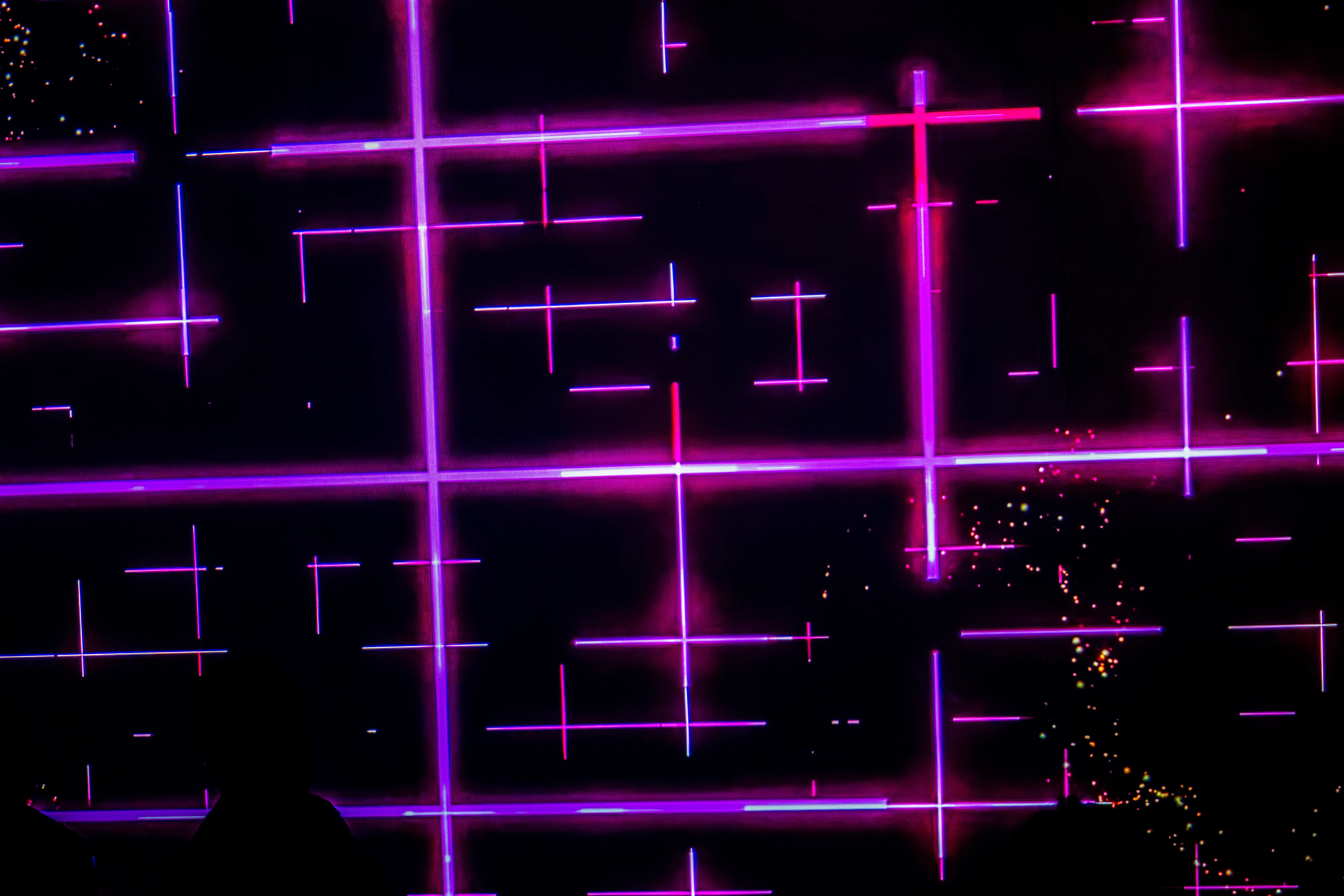 Silhouetted figures stand in front of a dark screen displaying a bright, abstract neon grid pattern in pink and purple, with scattered light particles.