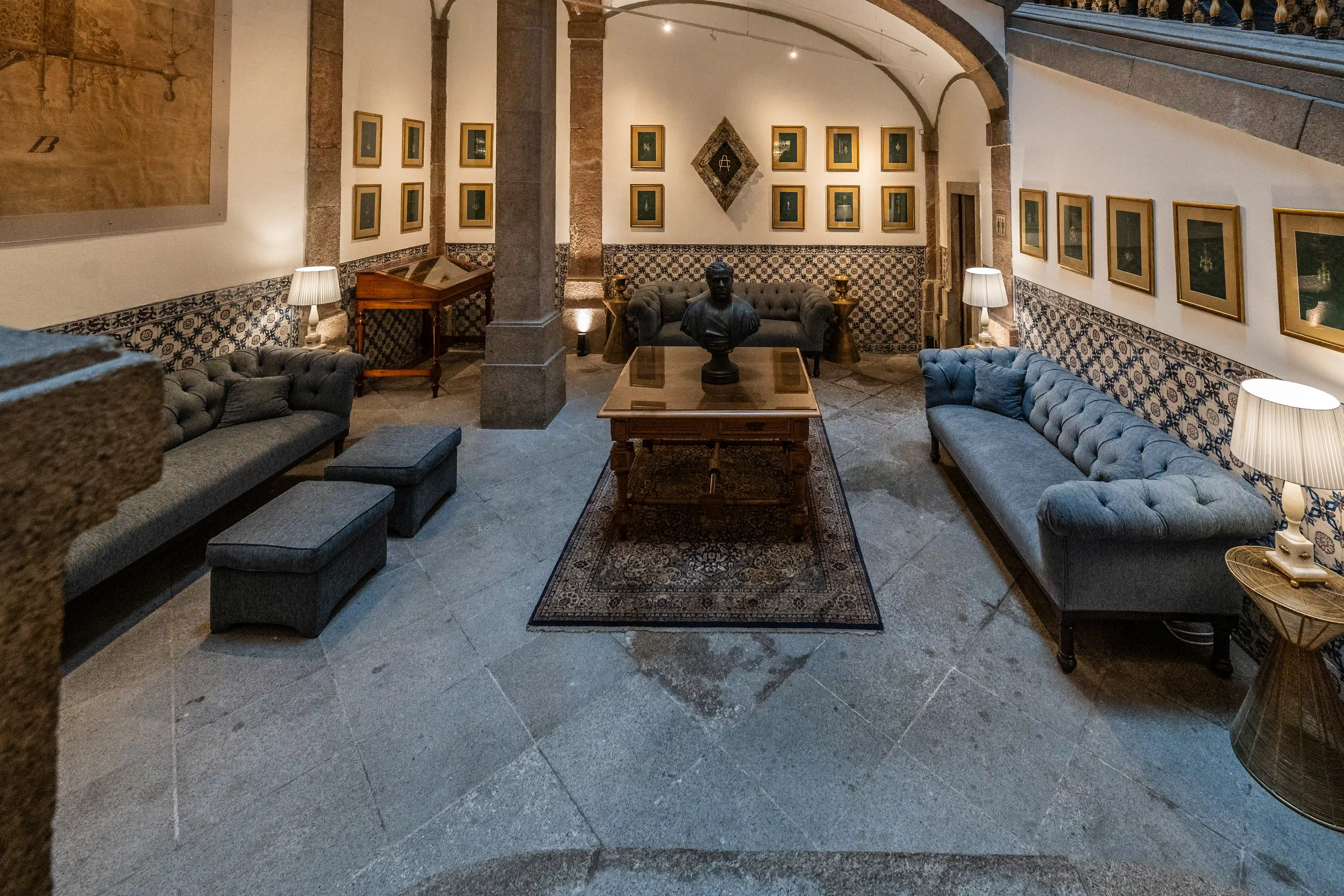A formal lounge area with two deep blue tufted sofas, a wooden coffee table with a bust, a patterned rug, and a tiled wall featuring framed artwork. Stone pillars and arched doorways add to the room's historic ambiance.
