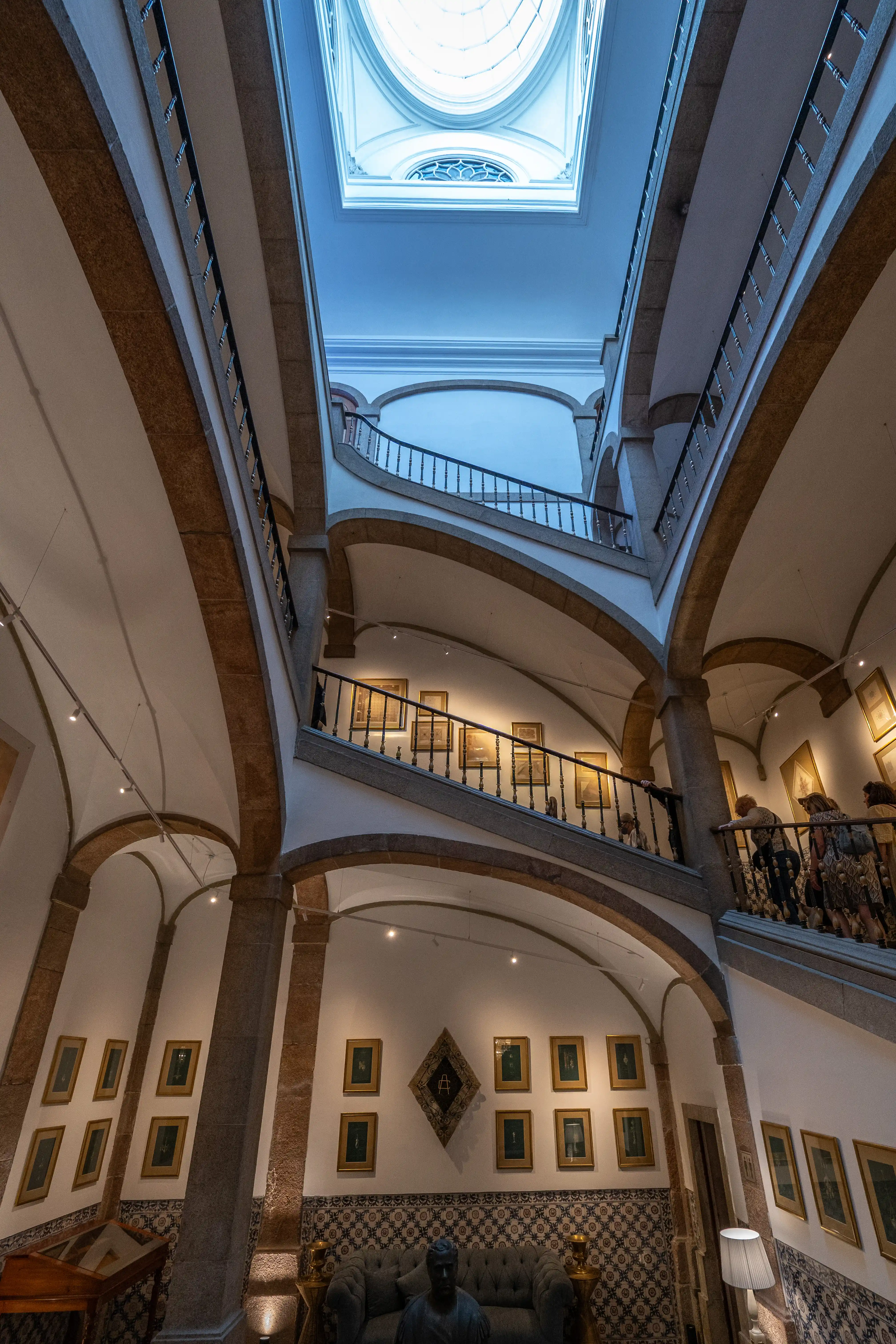 Wide-angle view looking up at a grand, multi-tiered staircase in a museum, featuring stone arches, a central skylight, framed artwork on the walls, and several visitors.