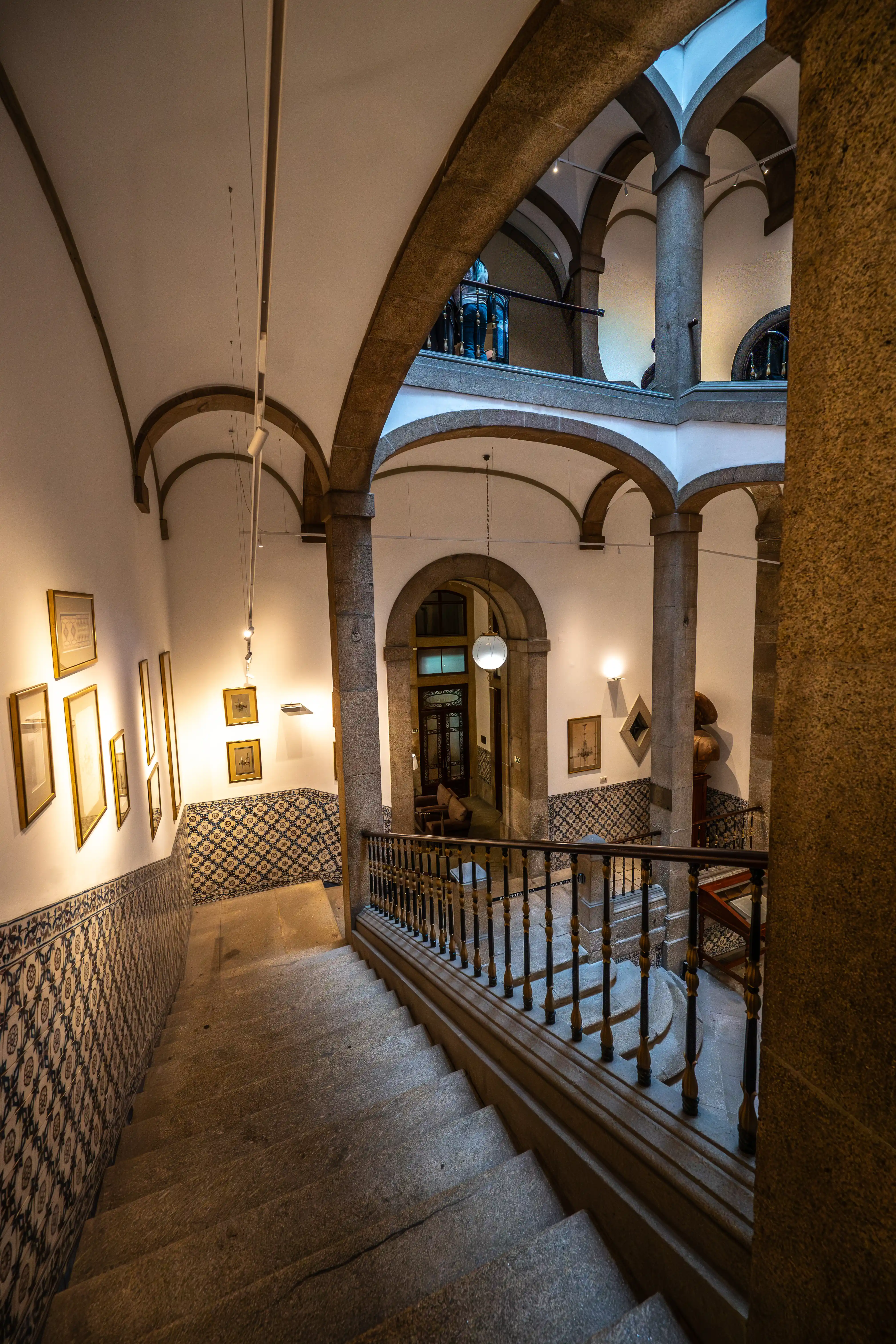 A wide stone staircase curves downwards, flanked by walls decorated with framed paintings and traditional blue and white tiled patterns. Arched stone columns and vaulted ceilings create a sense of grandeur in the interior space, with a decorative wooden banister guiding the way.