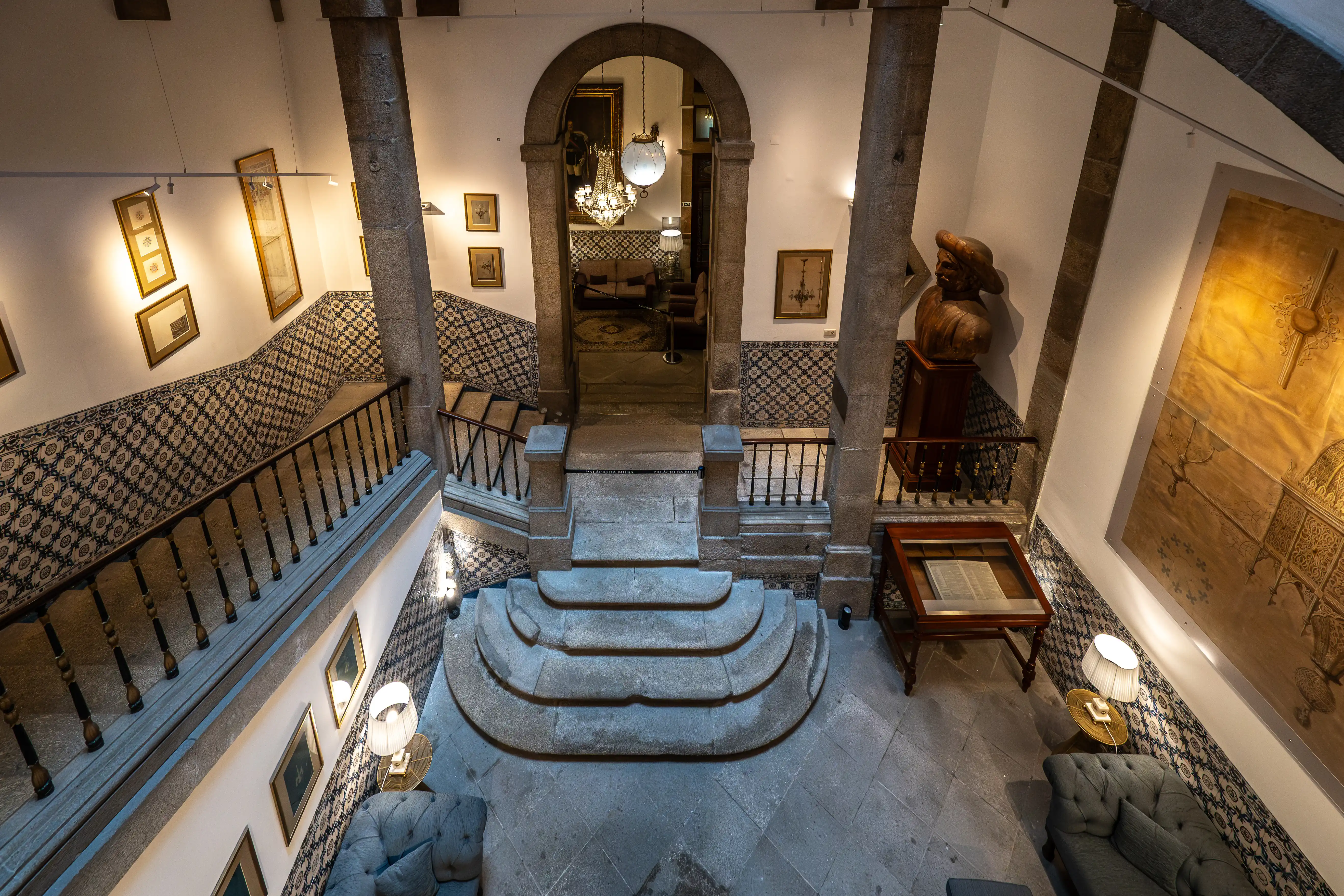  Interior view from above of a grand staircase hall with patterned tiles, stone steps, a bust, paintings, and antique furniture.