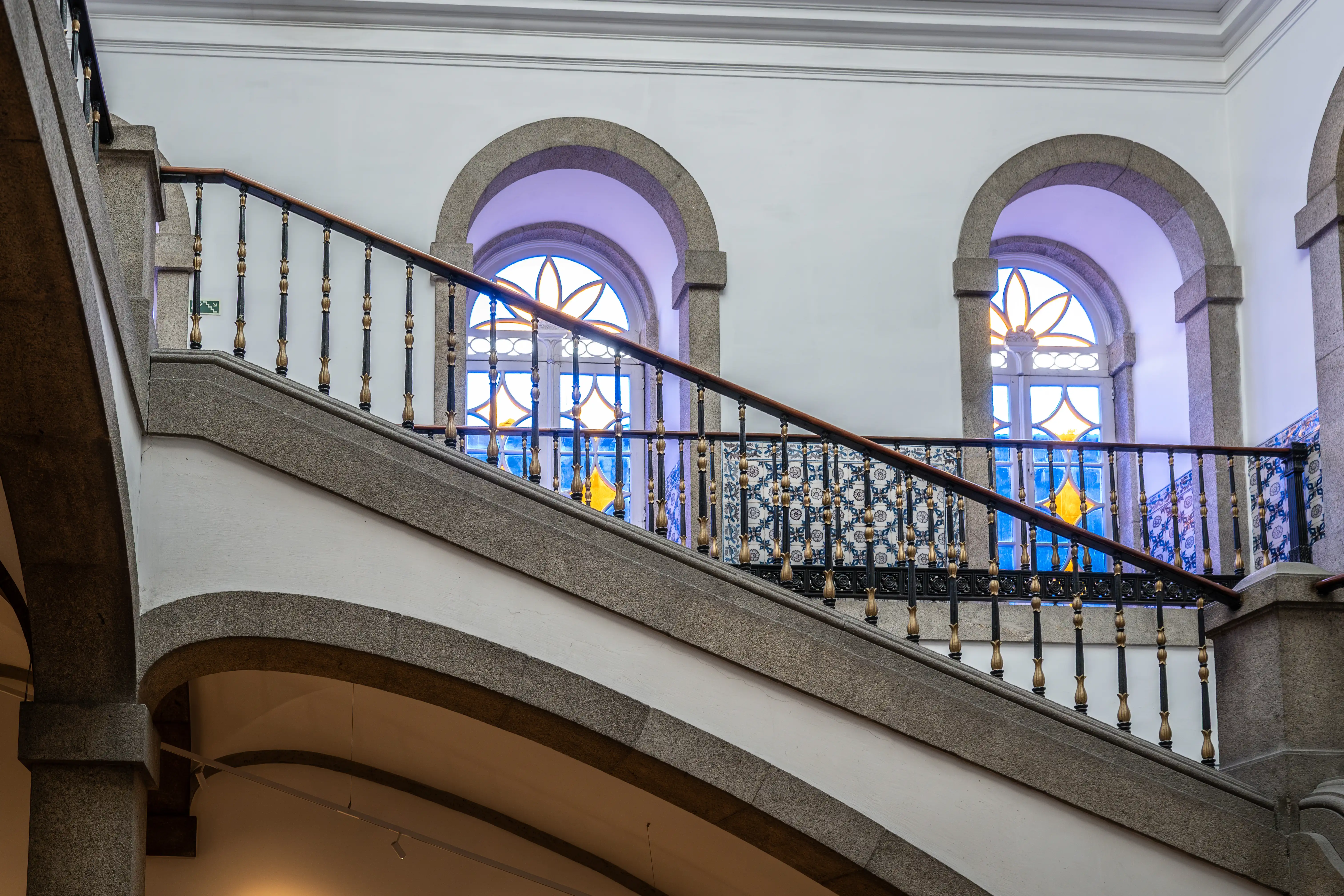 High-angle view of a grand interior staircase with curved stone steps, walls covered in black and white patterned azulejo tiles, framed artwork, a bust on a pedestal, a wooden banister, and a grey sofa with a lamp in the foreground.